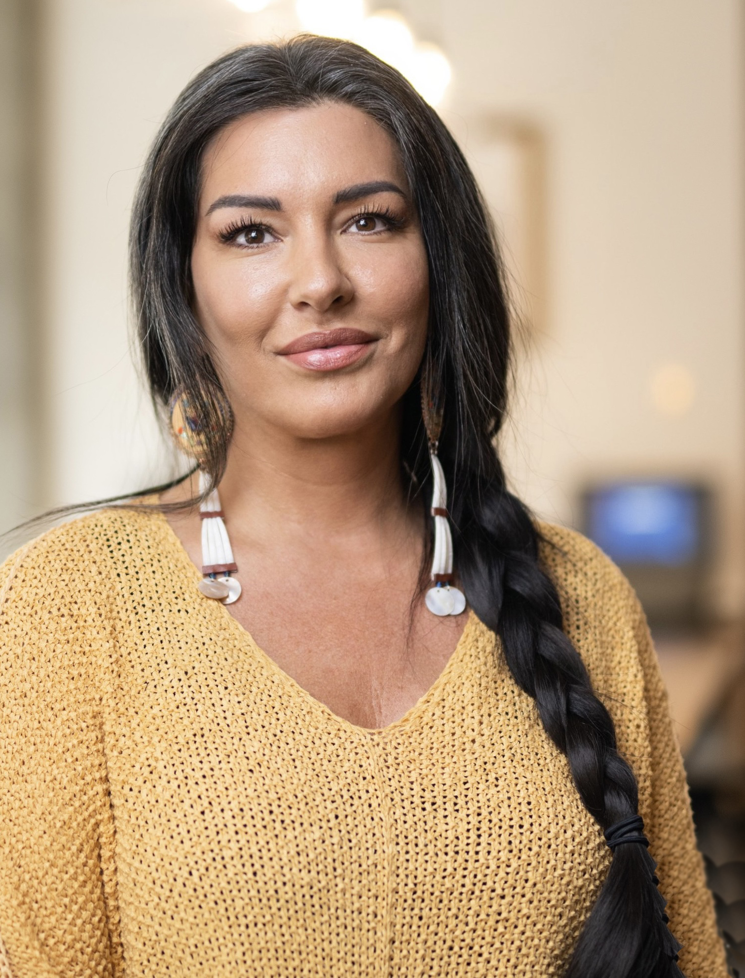 A woman with long black hair styled in a braid, wearing a yellow knit sweater and colorful earrings, standing indoors with a blurred background.