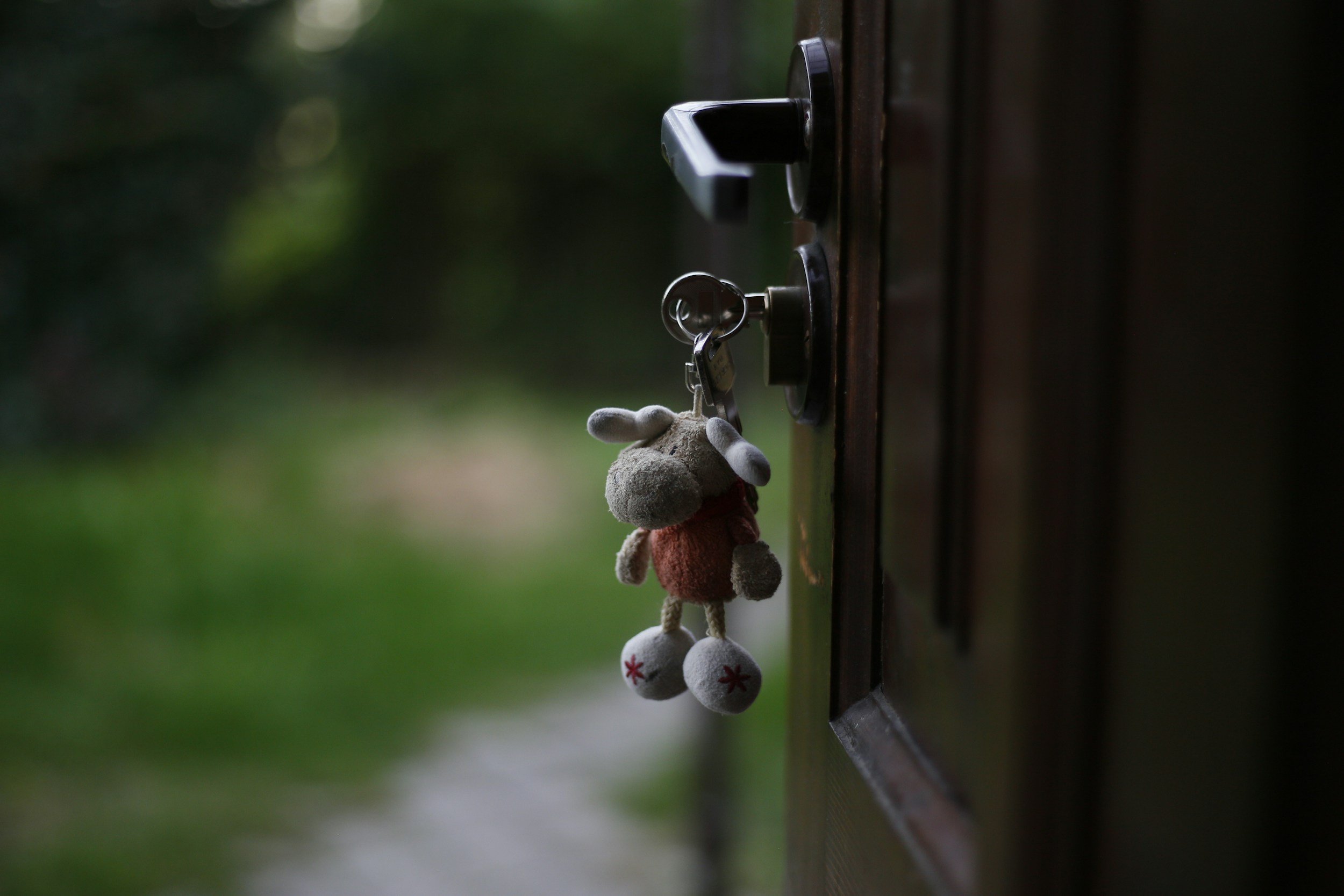 A plush toy hanging from a doorknob outside a wooden door, with blurred greenery in the background.