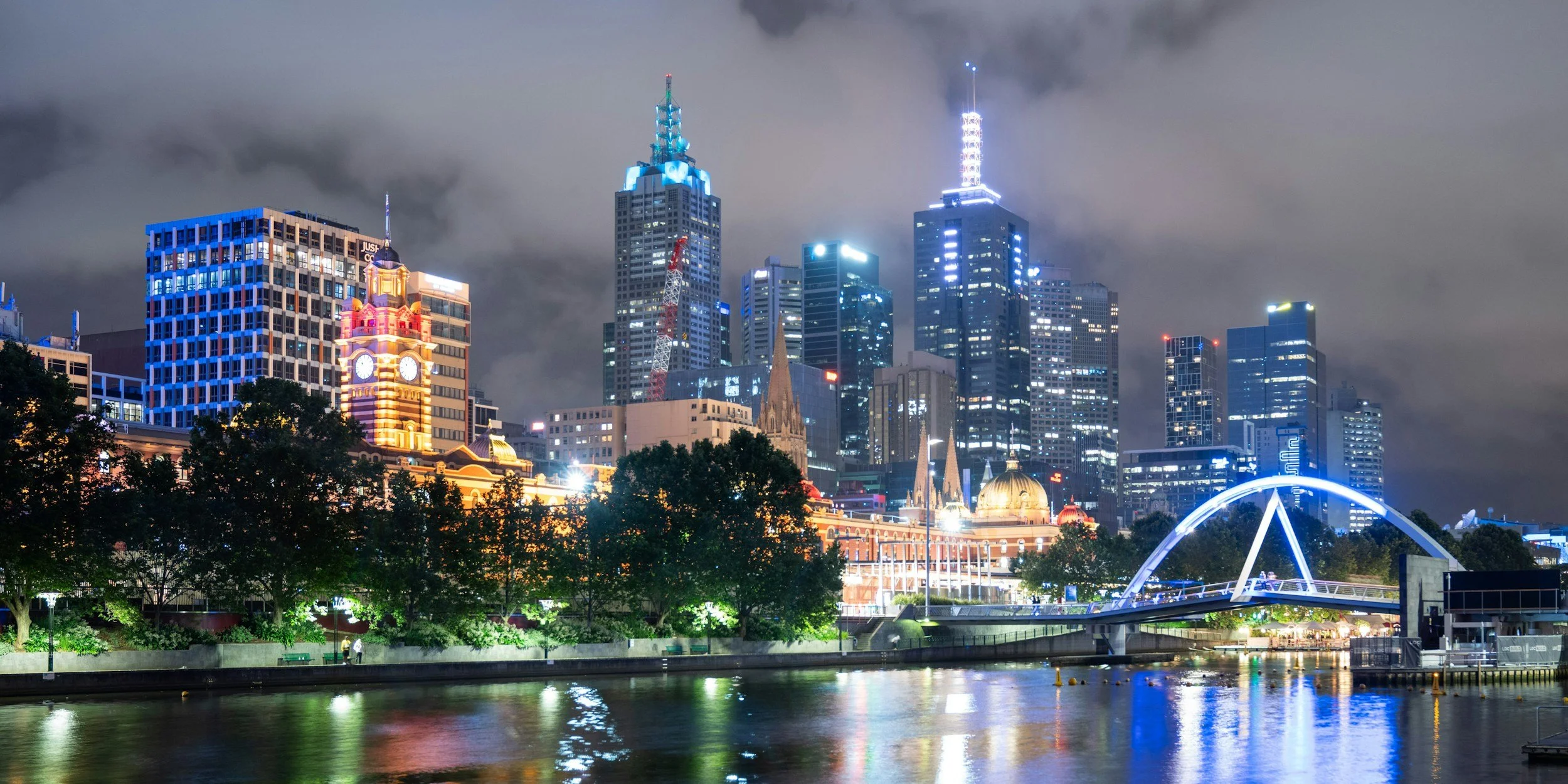 Night view of a city skyline with tall illuminated buildings, a bridge with bright blue lights over water, and a cloudy sky.