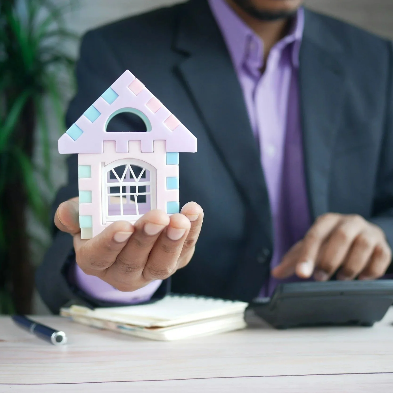 Person in a blazer holding a small colorful house model with a notepad and calculator on a desk.