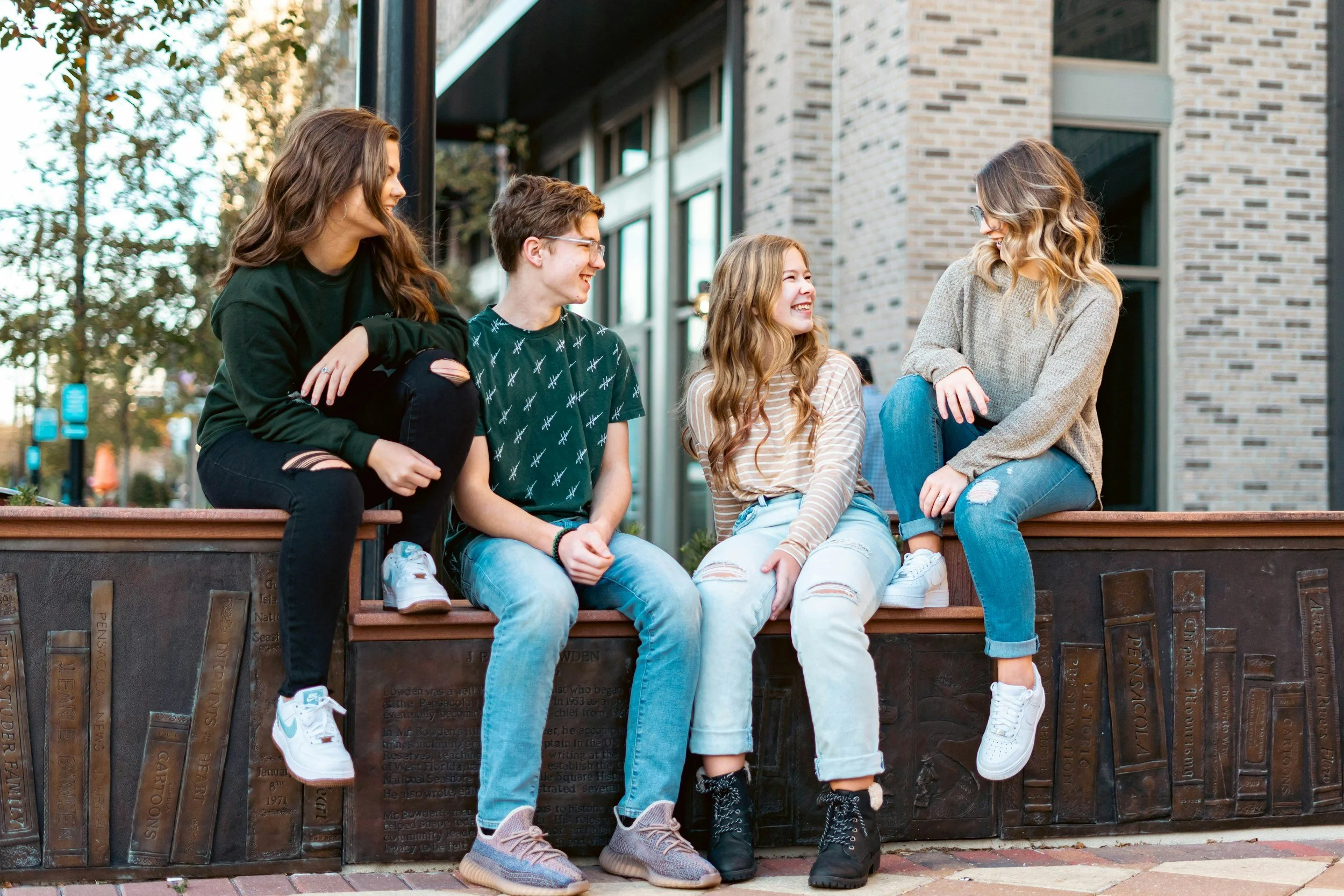 Four teenagers sitting on a low wall outside a modern brick building, smiling and engaging with each other on a sunny day.