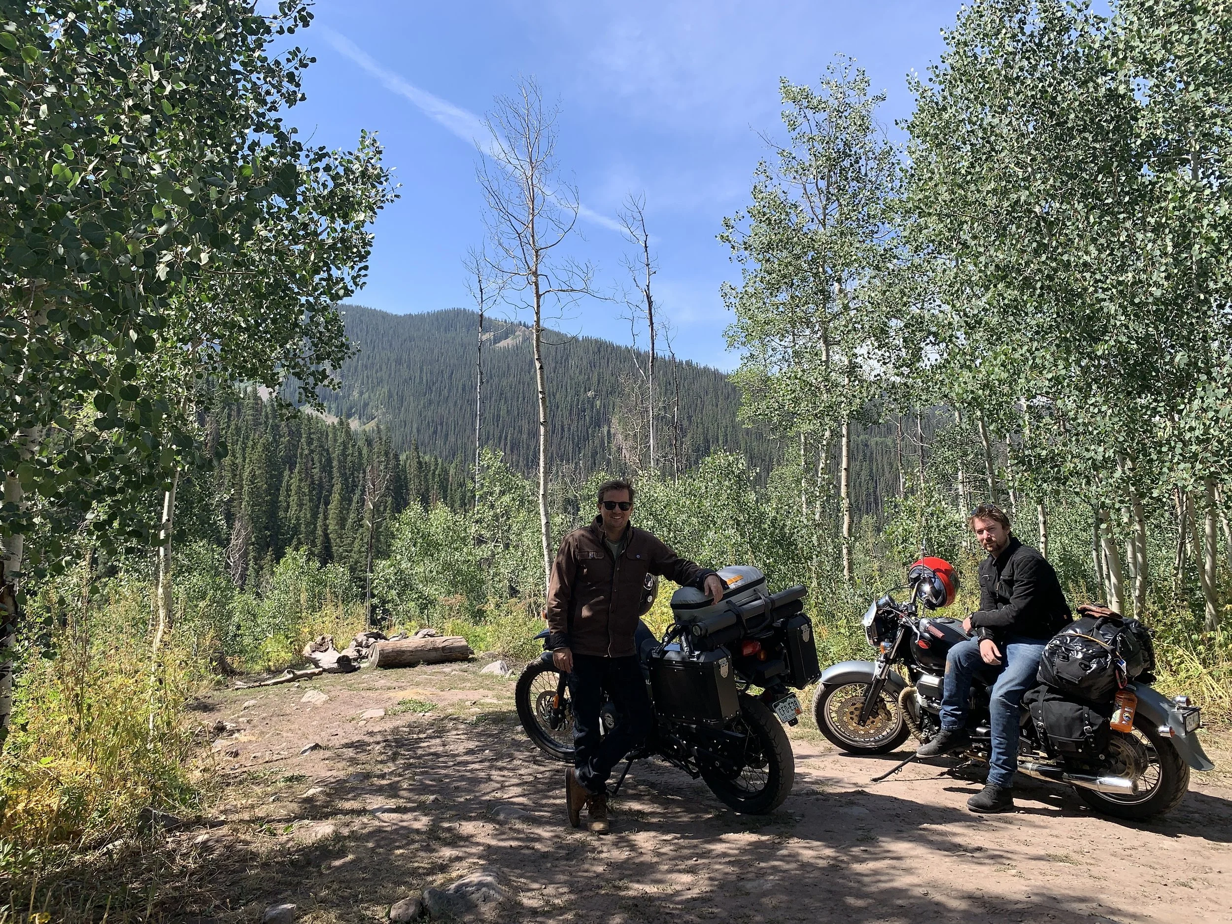 Two men with motorcycles on a dirt trail in a forested mountain area under a blue sky.