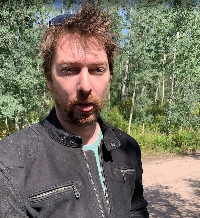 A man with brown messy hair and beard, wearing a black jacket, standing outdoors in a green, wooded area during daytime.