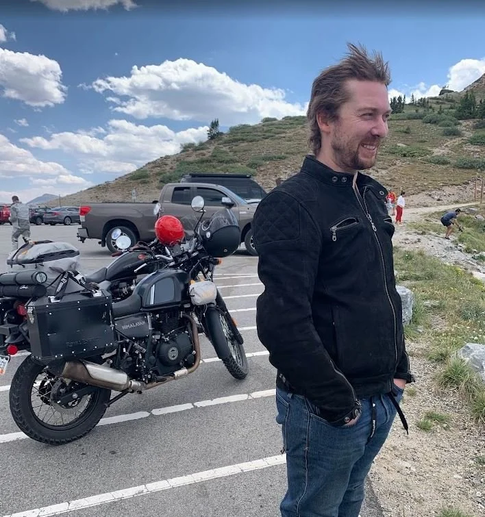 Man in black jacket smiling with his hands in his pockets, standing in a parking lot next to black motorcycle with saddlebags and helmets, with hills and a partly cloudy sky in the background.