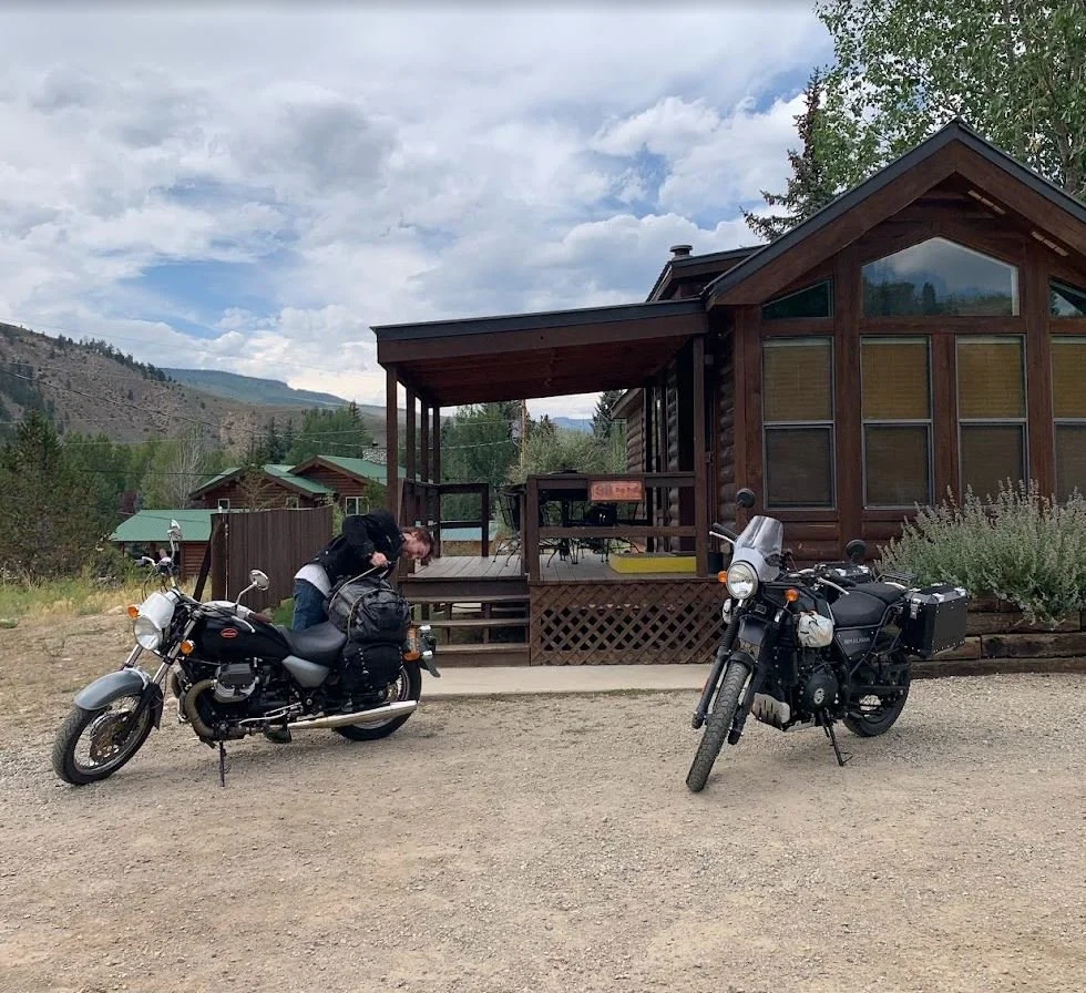 A person leaning over a black motorcycle with a helmet and backpack, parked outside a wooden cabin with a deck, in a mountainous area with trees and cloudy sky.