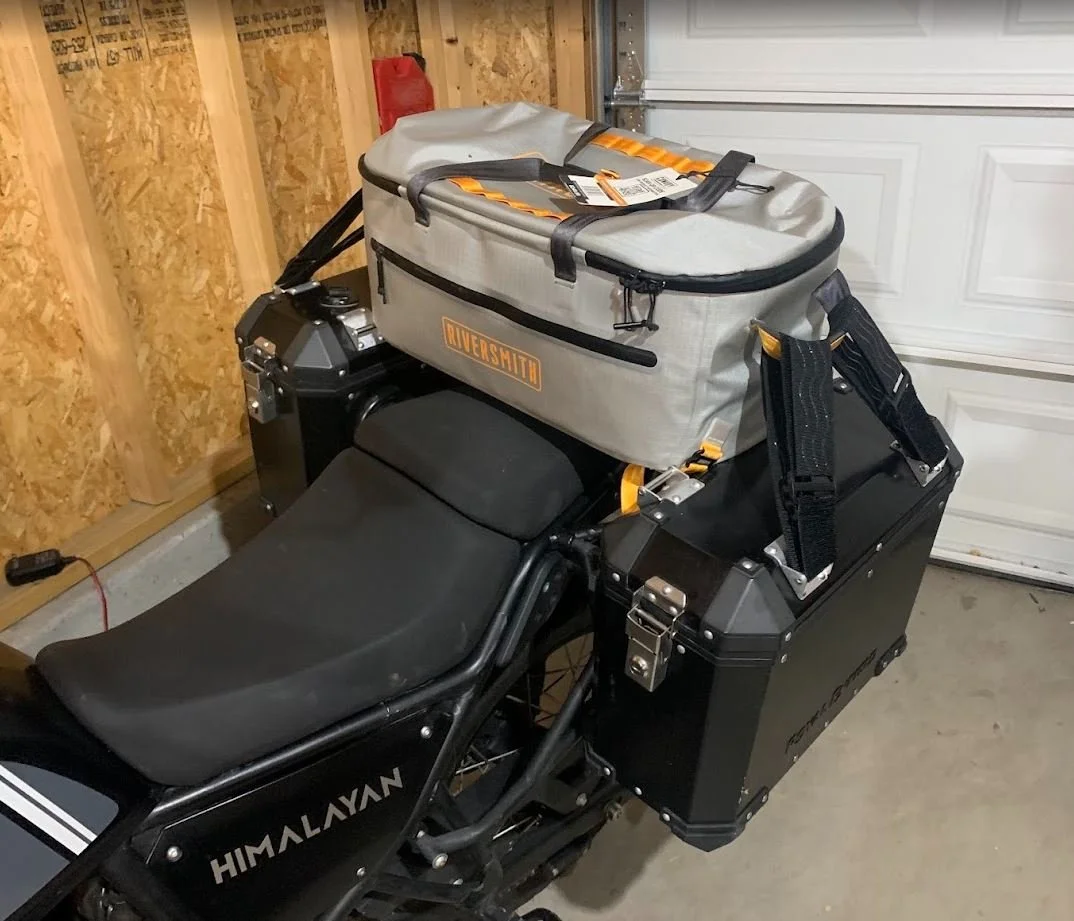A black Himalayan motorcycle with a gray Riversmith bag on top and a black side case, parked inside a garage with wooden framing and a closed white garage door.