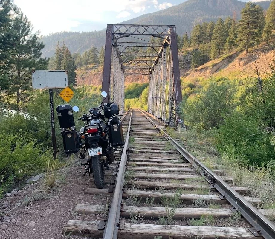 Motorcycle parked on a railway track leading to a bridge over a river, with a sign on the left that says "Keep Left of Bridge." The area is surrounded by trees and green hills with mountains in the background.