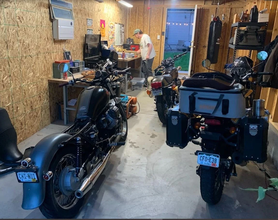 A garage with three motorcycles and a person working at a workbench, with tools and equipment on shelves and a door leading outside.
