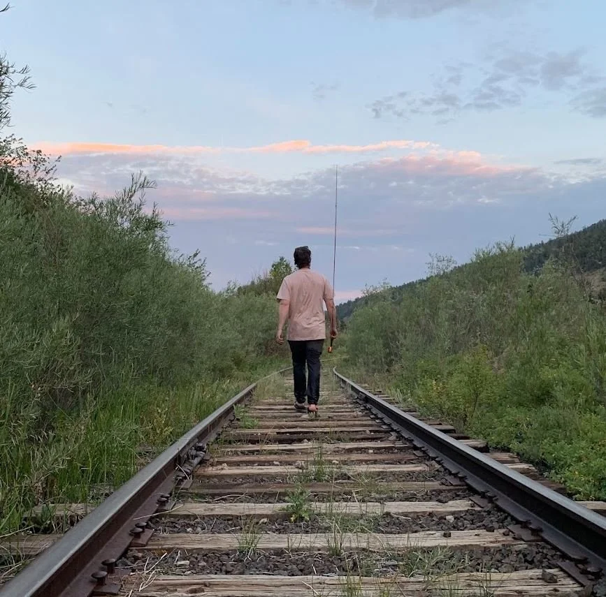 A person walking on train tracks through a green, hilly landscape at dusk, holding a fishing rod.