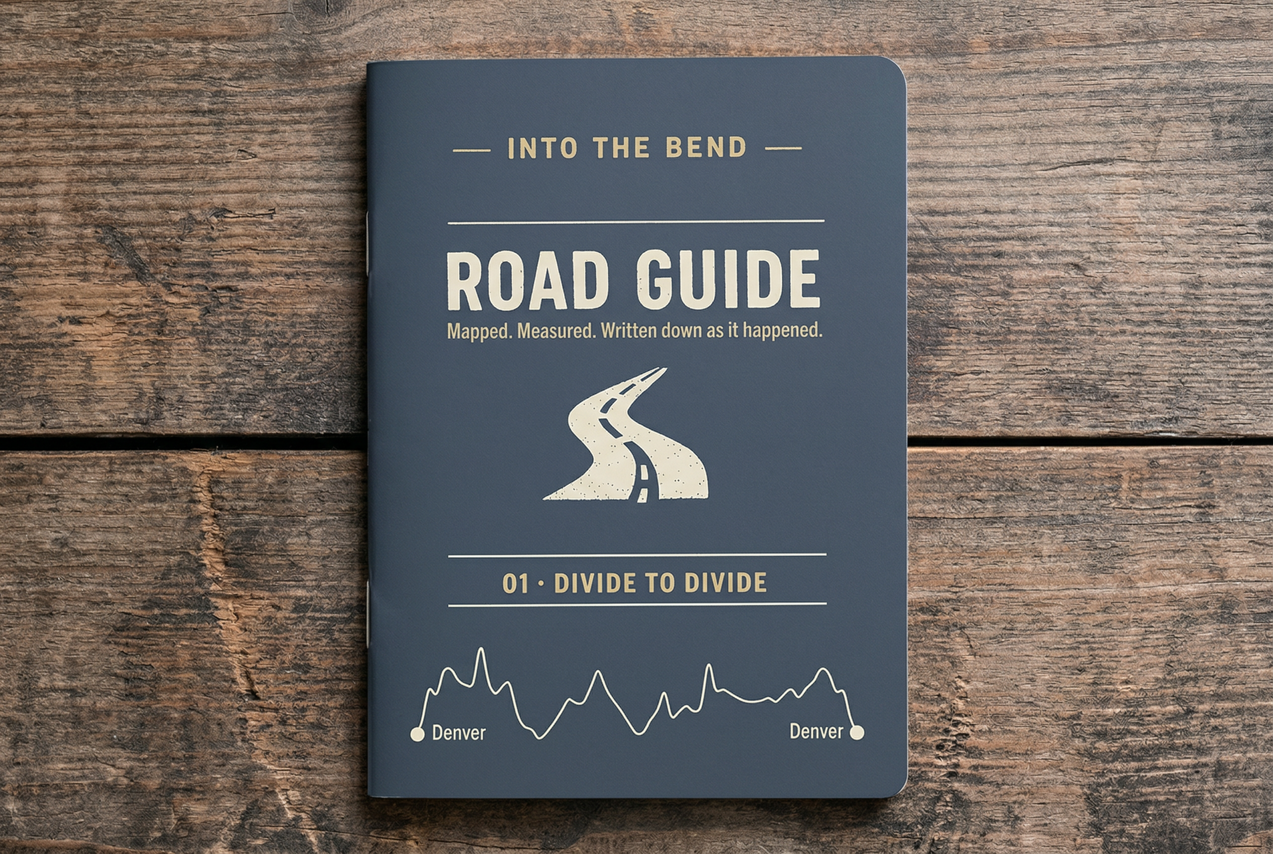 A dark blue road guide notebook titled "Into the Bend Road Guide" on a wooden surface, with a graphic of a winding road and the phrase "Mapped. Measured. Written down as it happened." Additionally, it has a section labeled "01 - Divide to Divide" with a simple line graph showing a fluctuating route between Denver and Denver.