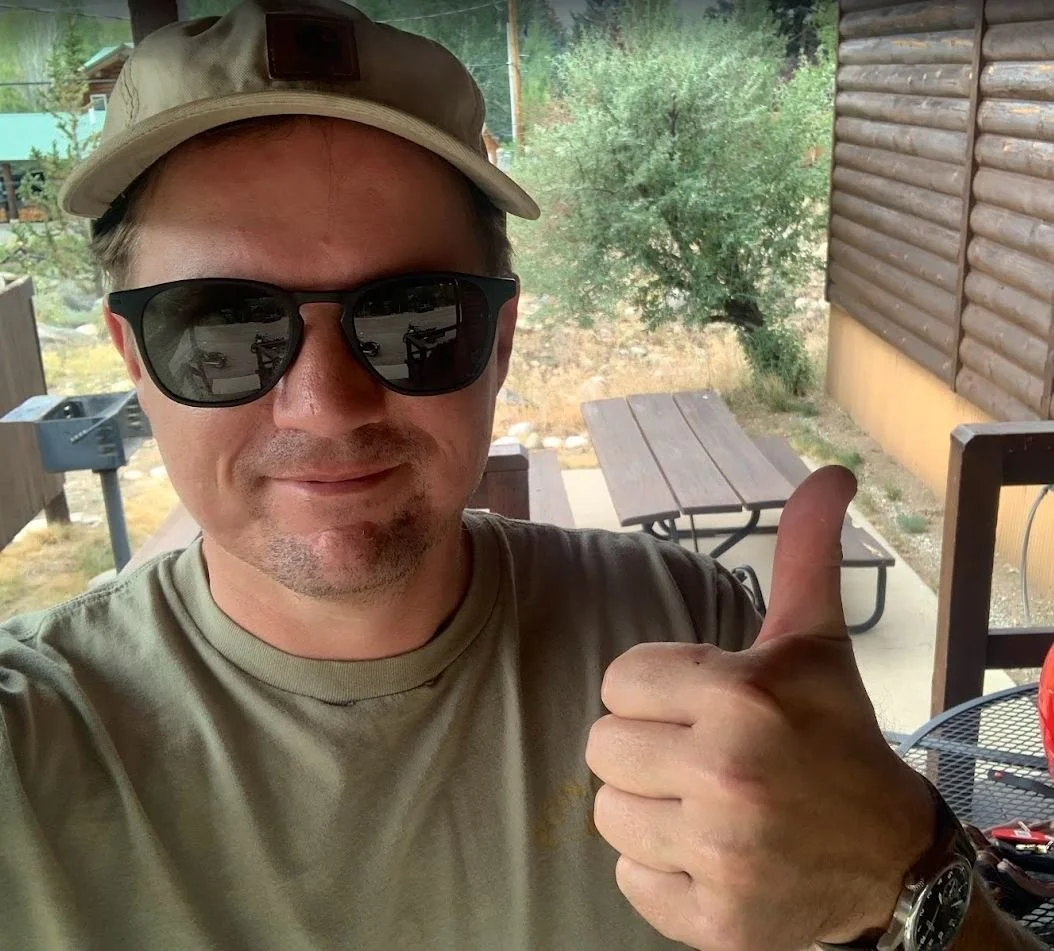 A man wearing a tan cap, black sunglasses, and a beige t-shirt giving a thumbs-up while smiling outdoors on a patio.