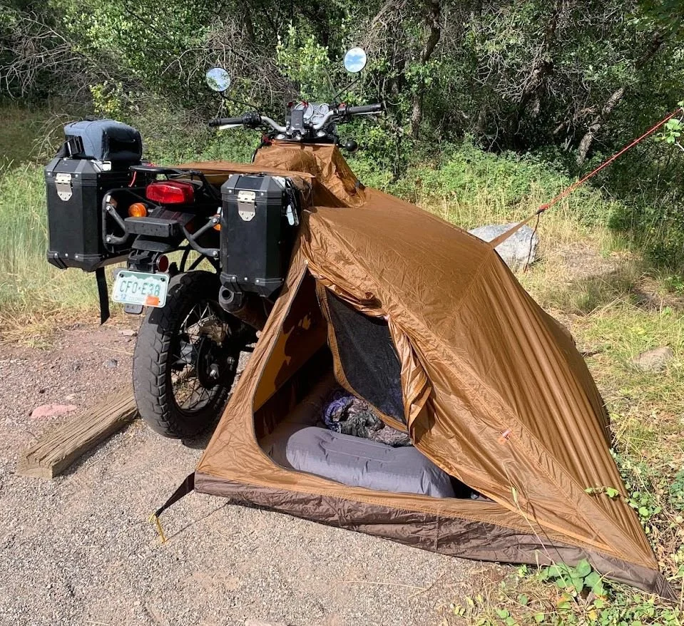 A motorcycle with a brown rain cover set up as a tent, with a sleeping bag inside, on a dirt path in a wooded area.