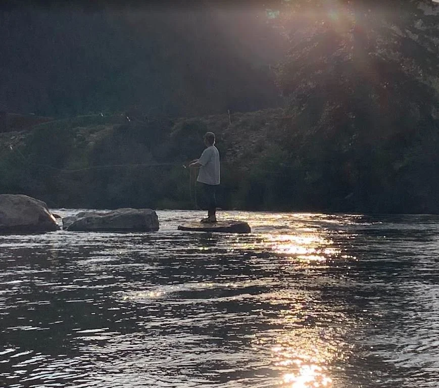 A person standing on a rock in a river, fishing with a rod during sunset.