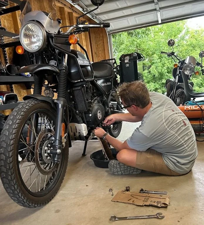 A man repairing a black Royal Enfield motorcycle in a garage, surrounded by tools and another motorcycle in the background.