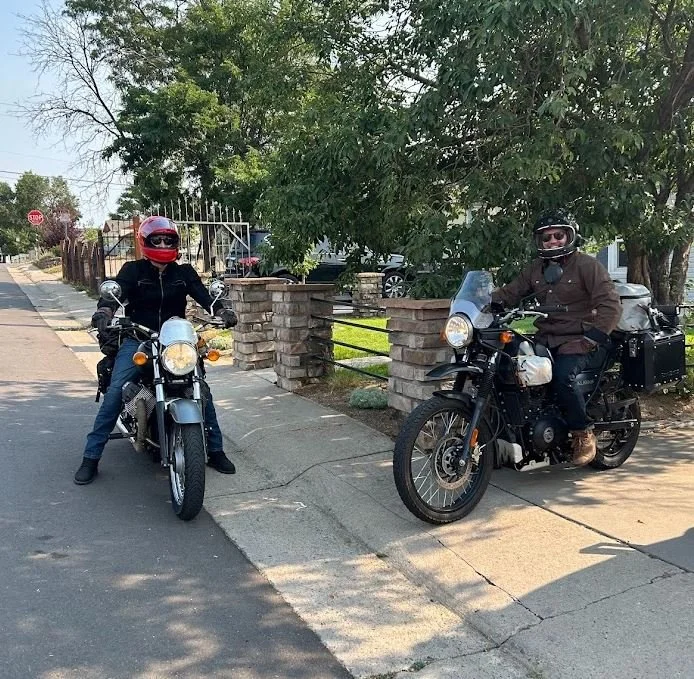 Two people sitting on motorcycles parked on the sidewalk near a stone fence with a large tree in the background.