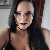 A woman with long black hair, wearing glasses, dark lipstick, and a black choker, taking a selfie indoors.