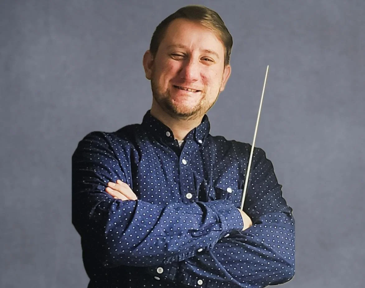 Man smiling with arms crossed, wearing a blue polka dot shirt, holding a conductor's baton, standing against a gray background.
