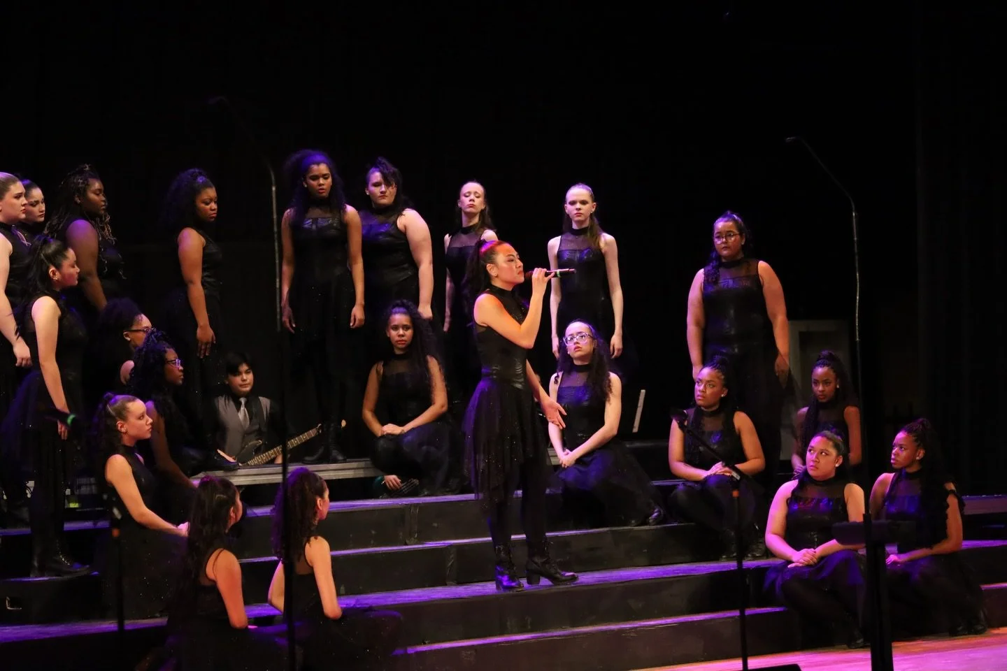 A group of young girls dressed in black on a stage, some sitting and some standing, with one girl in the center singing into a microphone and another girl playing a guitar.