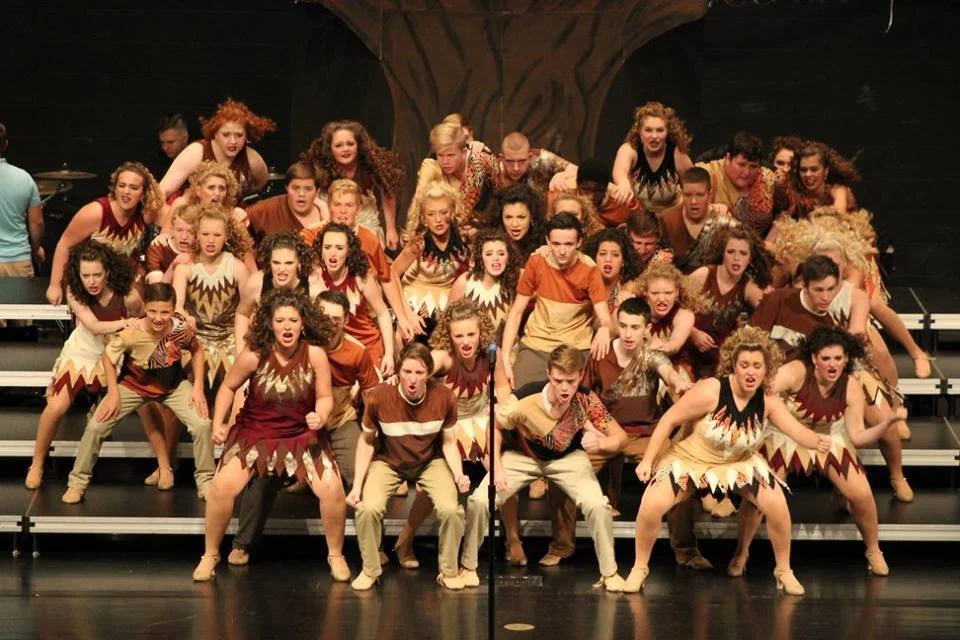 Group of performers on stage, dressed in tribal-themed costumes, performing a dance or musical number in a theater.