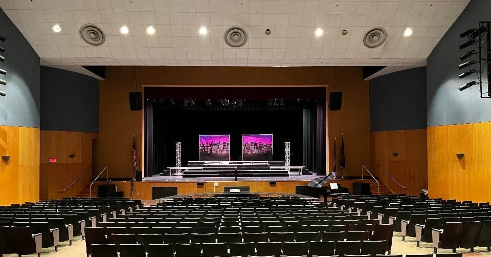 Empty indoor auditorium with a stage, two large screens displaying a cityscape, black chairs arranged in rows, and flags on either side of the stage.