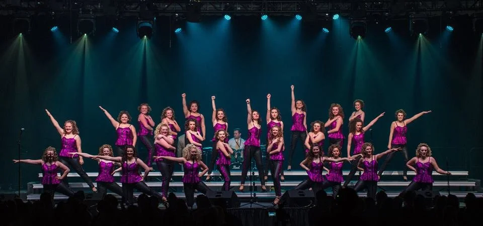 Group of young girls in purple costumes performing a dance on stage with stage lights and band members in the background.