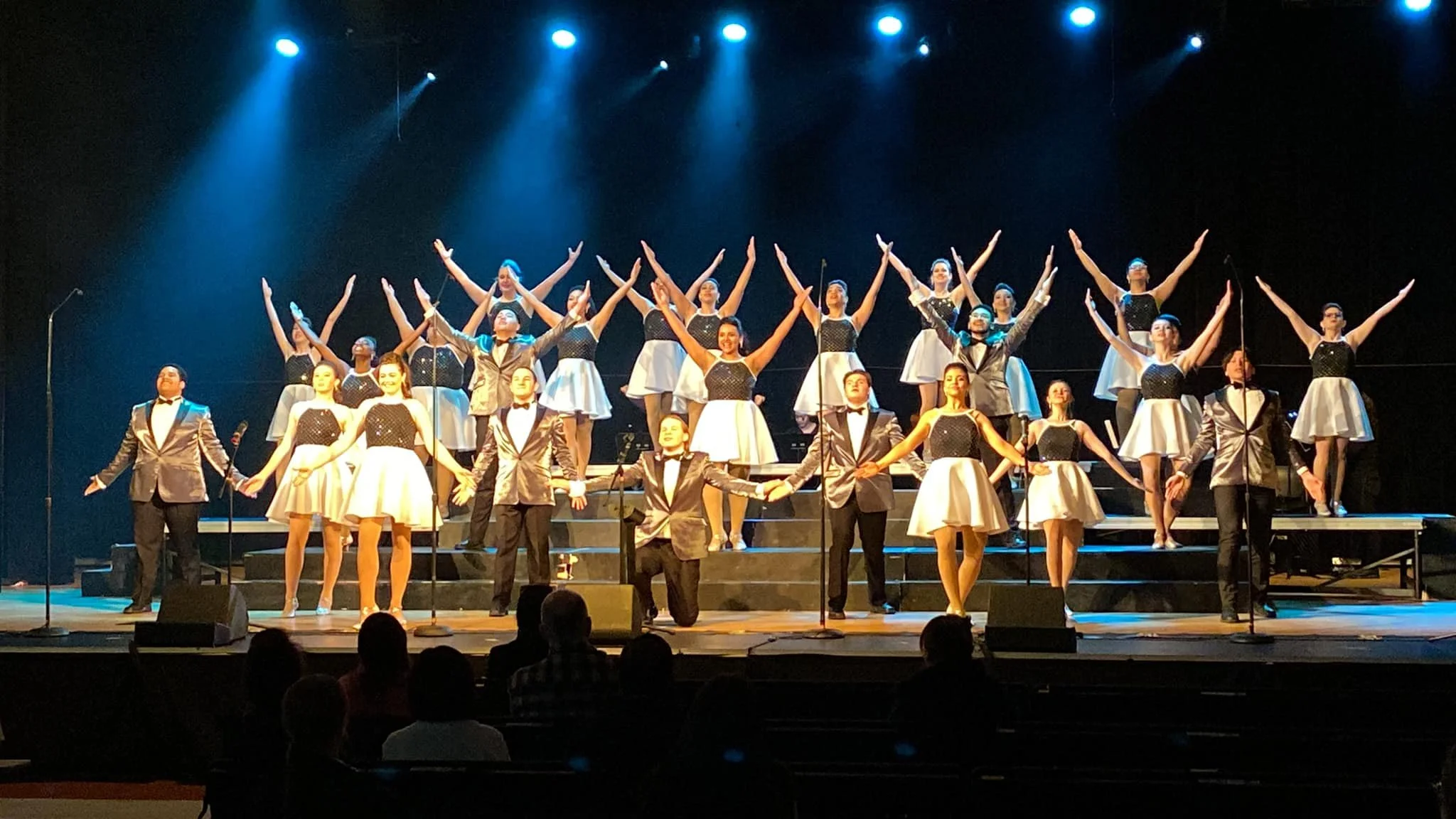 A large group of performers, both men and women, on stage during a theatrical or musical performance, dressed in formal attire. The women are in black and white dresses, and the men are in tuxedos. They are arranged on tiered platforms, with some standing, some kneeling, and some with arms outstretched, under stage lights.