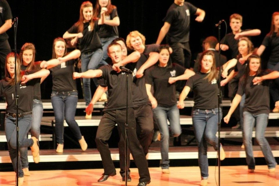 Group of teenagers performing a choreographed dance on stage, wearing matching black T-shirts and jeans.