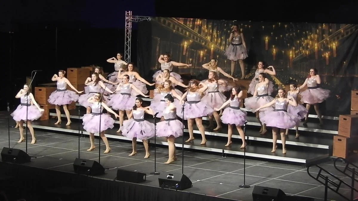 A dance performance on stage with dancers in purple tutus and silver tops standing on risers practicing a dance routine.