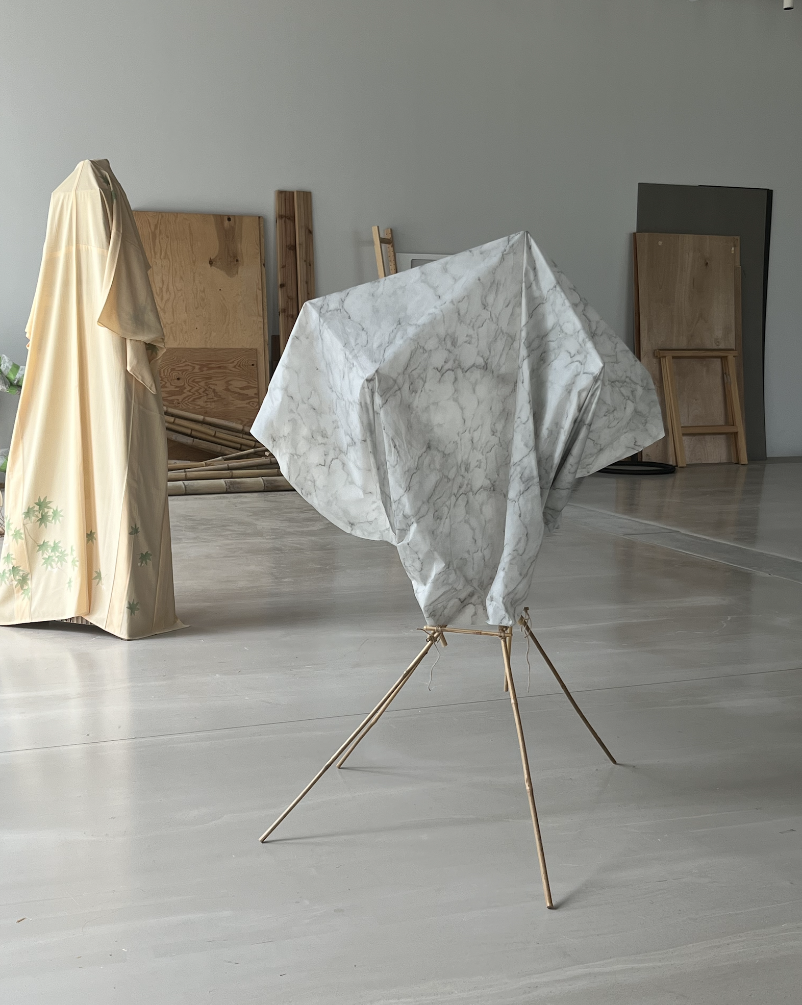 An art installation with a tripod stand and a marble-patterned fabric draped over it, resembling a sculpture in a gallery space with wooden and dark panels in the background.
