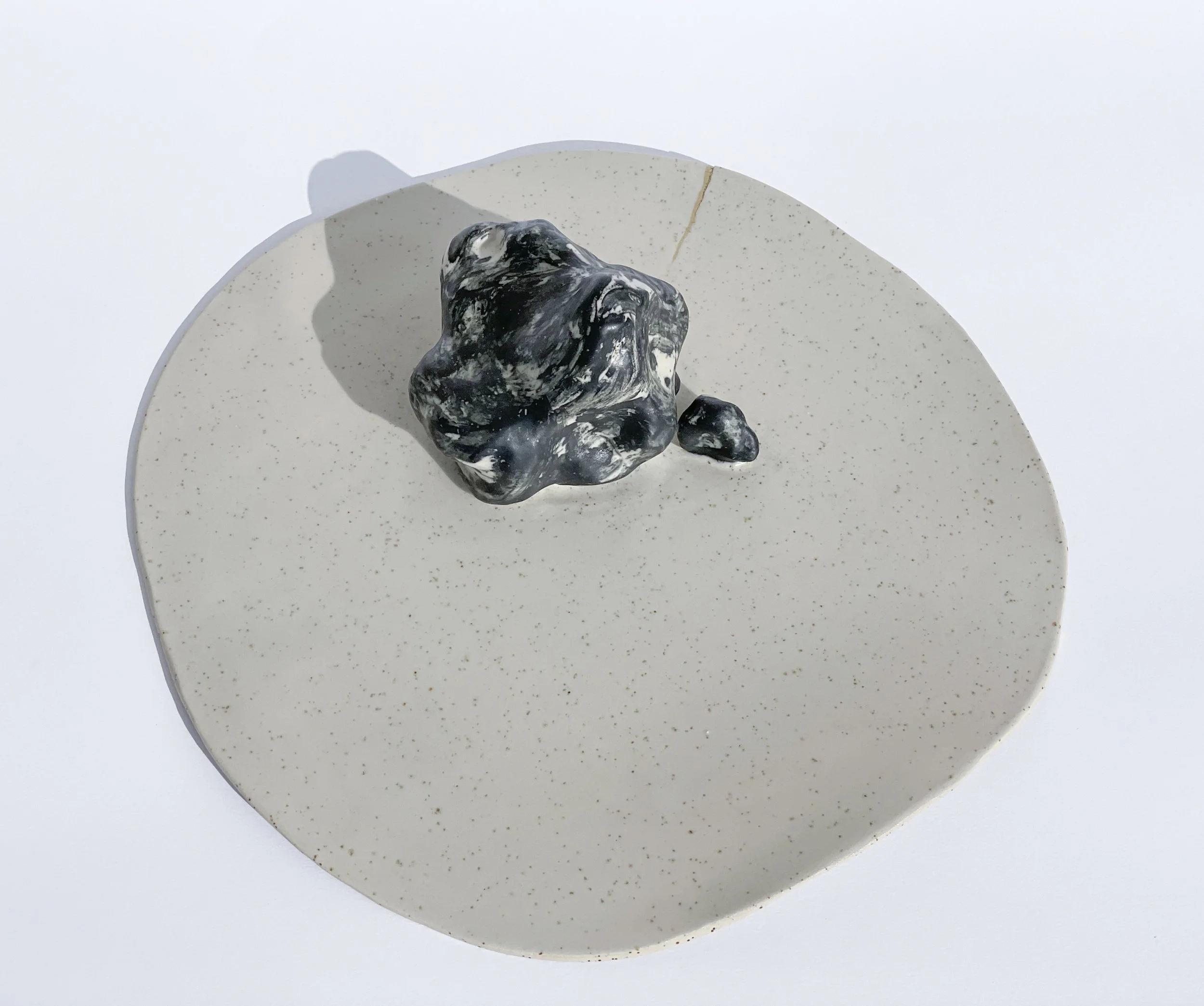 A black and white marbled sculpture on a round, speckled beige platform, casting a shadow on a white background.