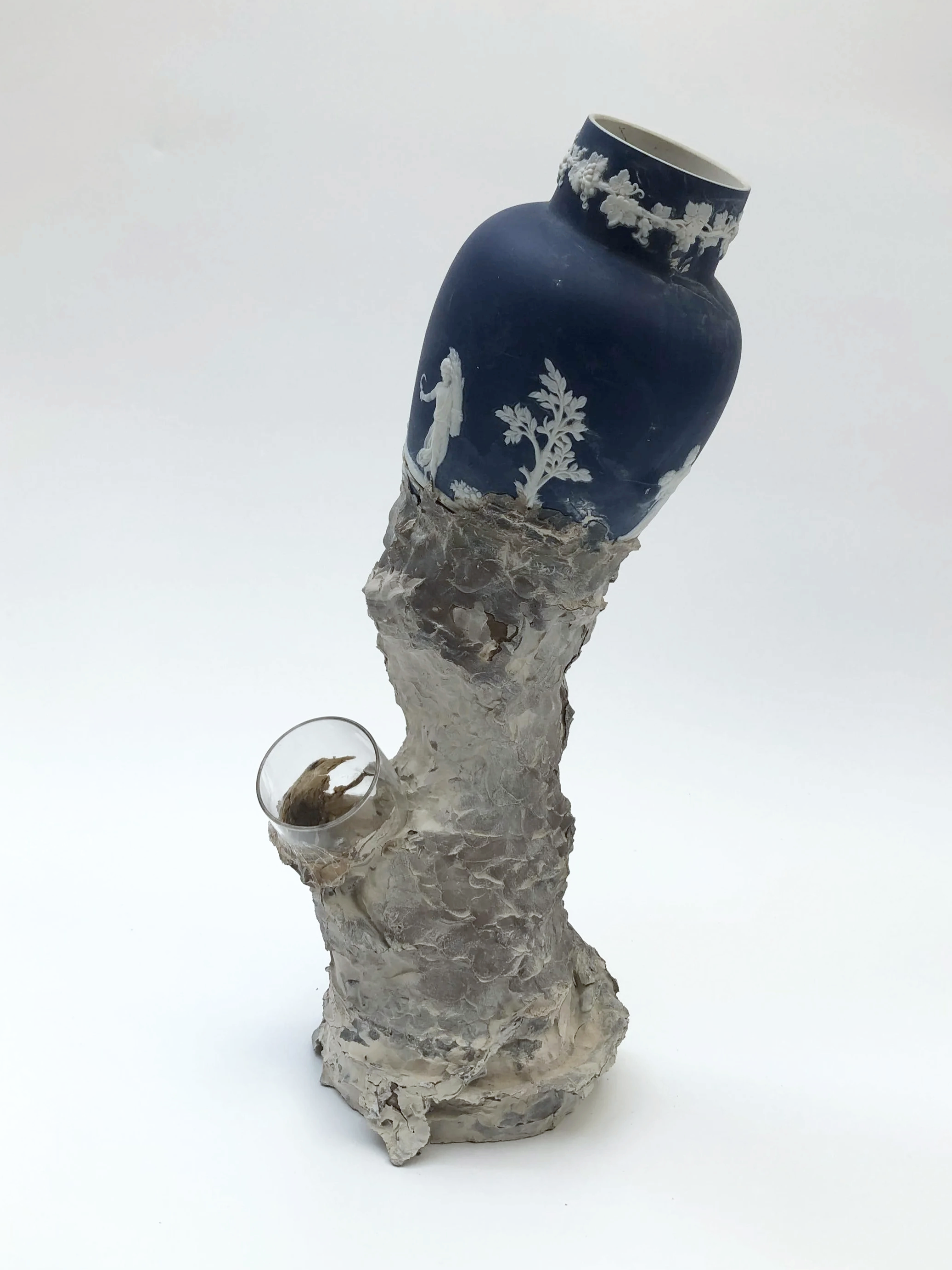 A sculpture of a blue and white porcelain vase with a tree and figure design, mounted on a textured, irregular, pale-colored base with a glass cup holding a small object, set against a plain white background.