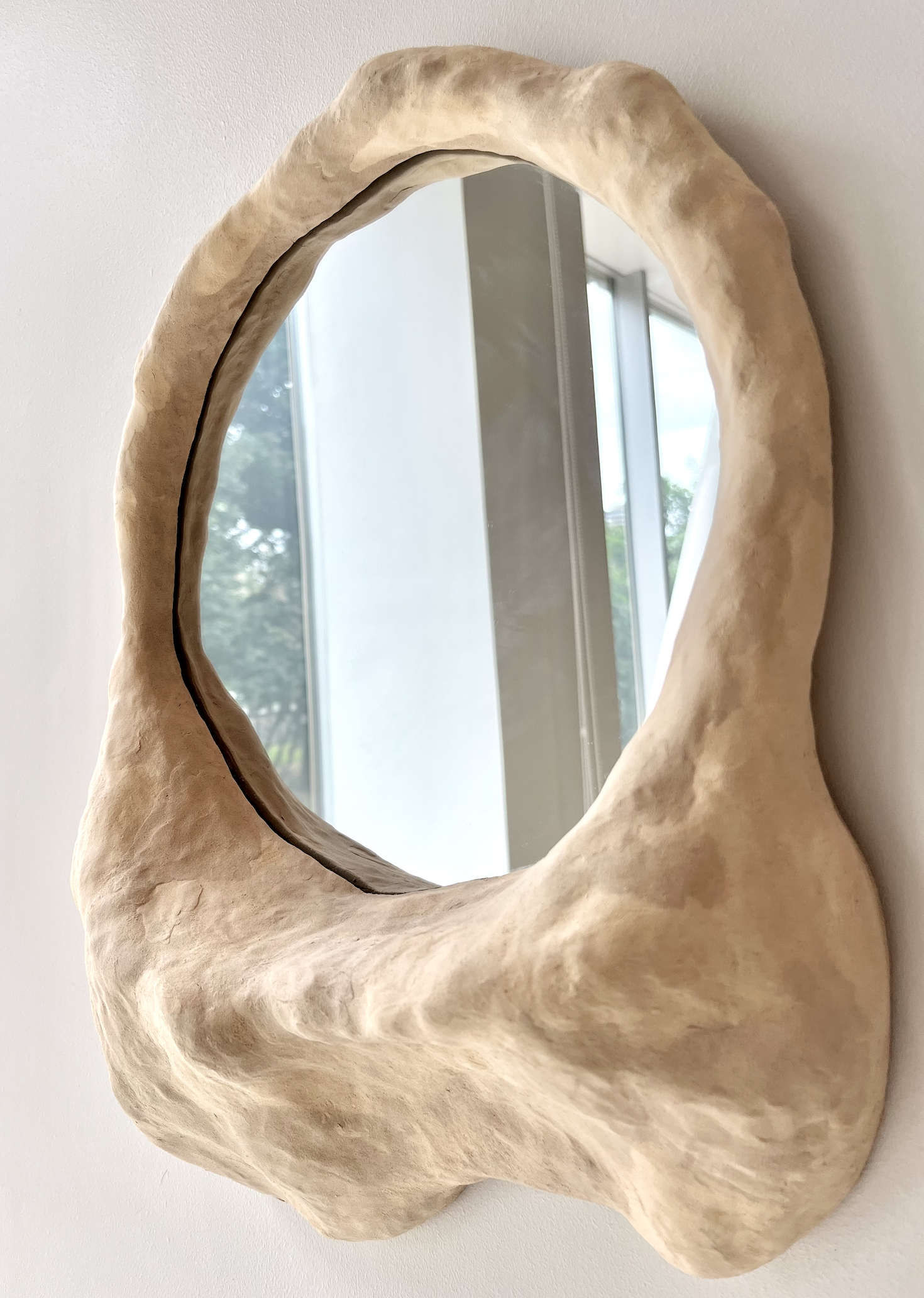 A decorative mirror with a thick, irregular,, beige-colored frame resembling a natural, organic shape, mounted on a white wall.