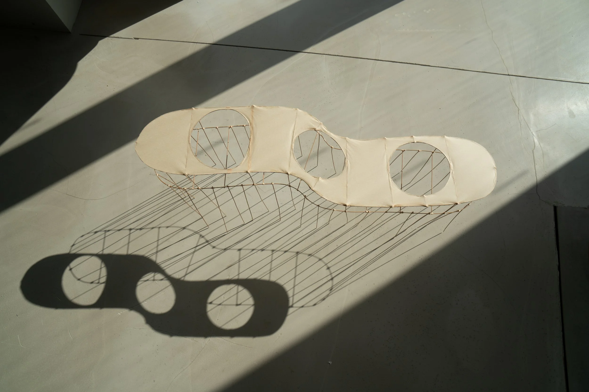 A modern art sculpture with an elongated, abstract shape made of fabric and thin metal rods, casting a shadow on a concrete floor.