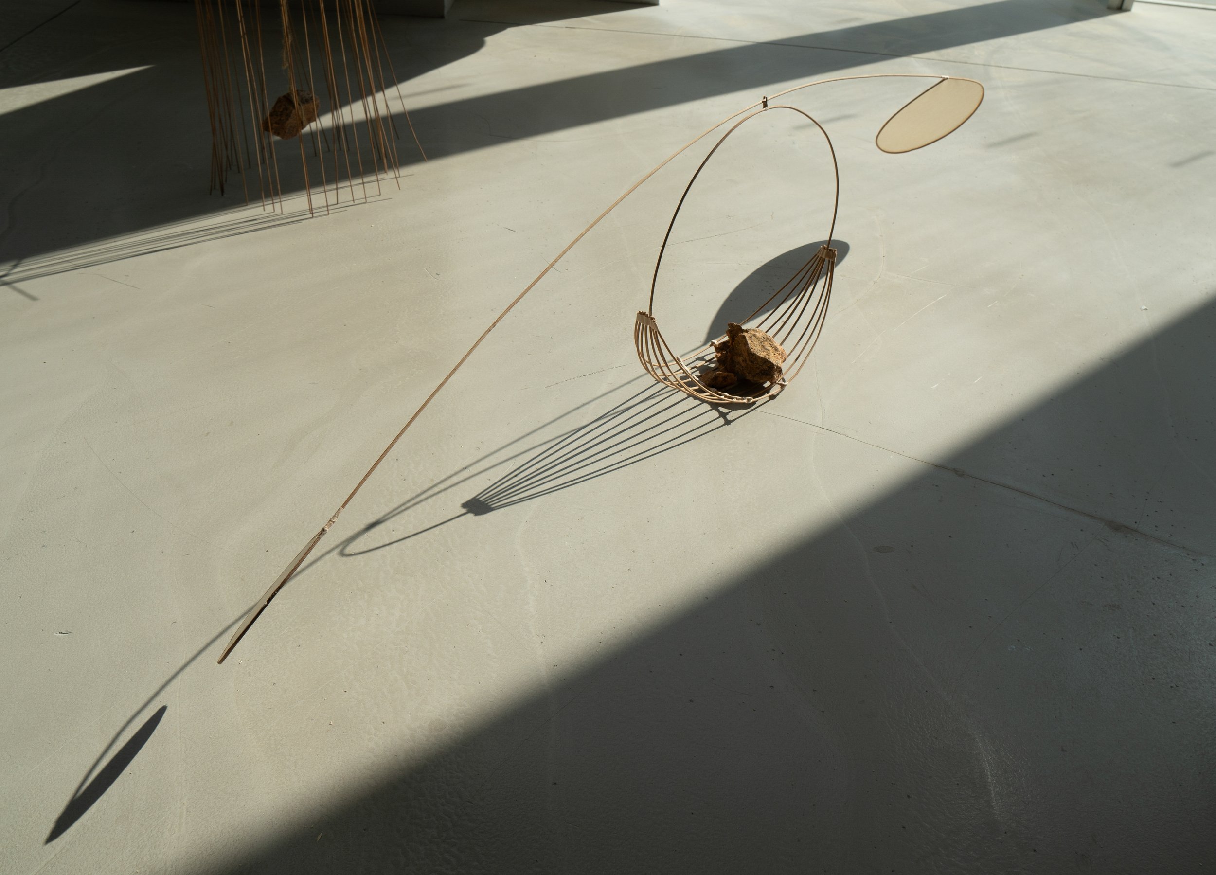 A minimalist art installation of a delicate sculpture with thin, curved metal rods and a suspended circular disk, casting long shadows on a light-colored floor.