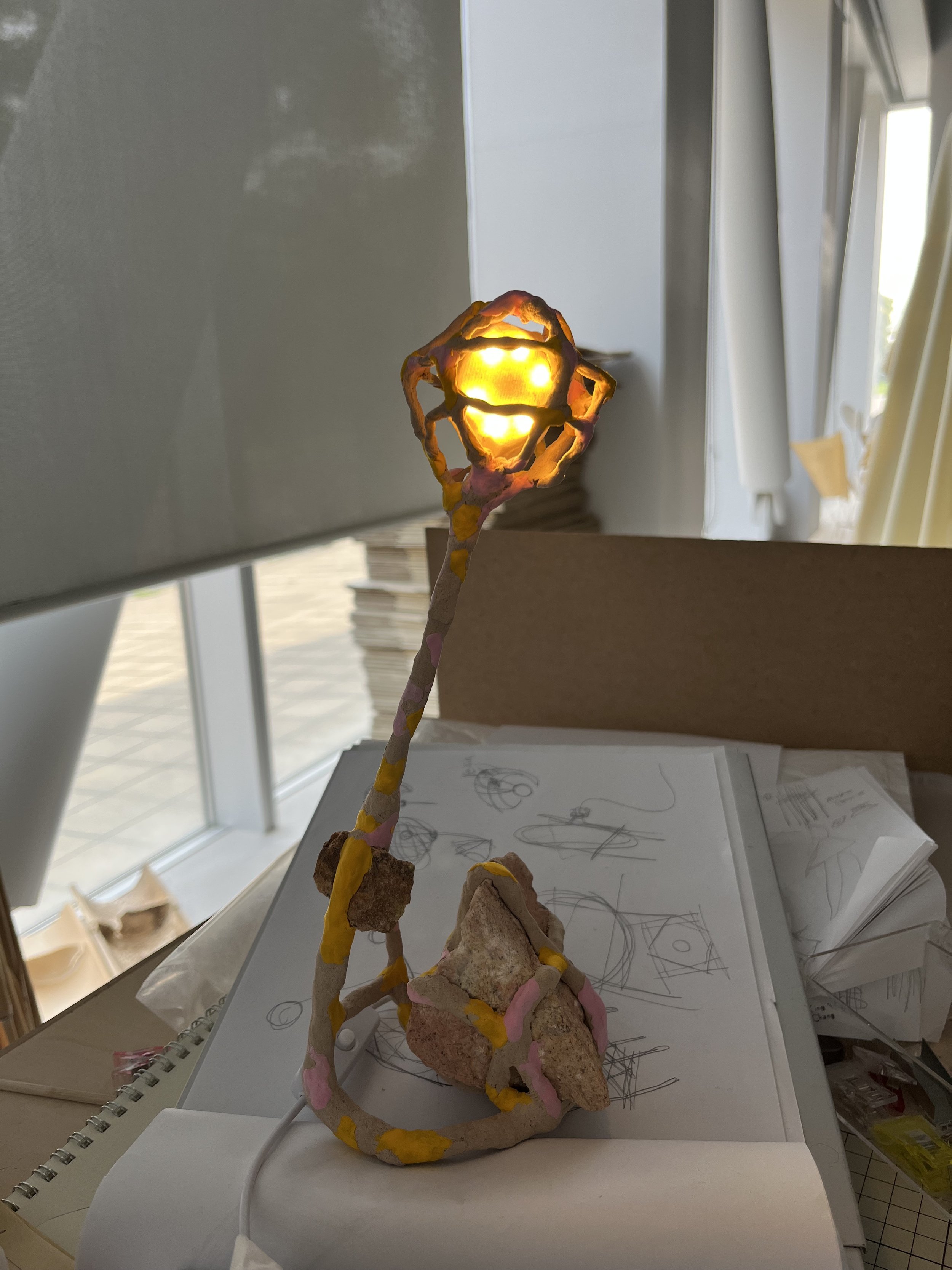 A handmade lamp resembling a flower with a glowing light bulb in the center, made from painted rocks and wrapped with colorful paper, placed on a desk with sketches and papers in the background near a window.