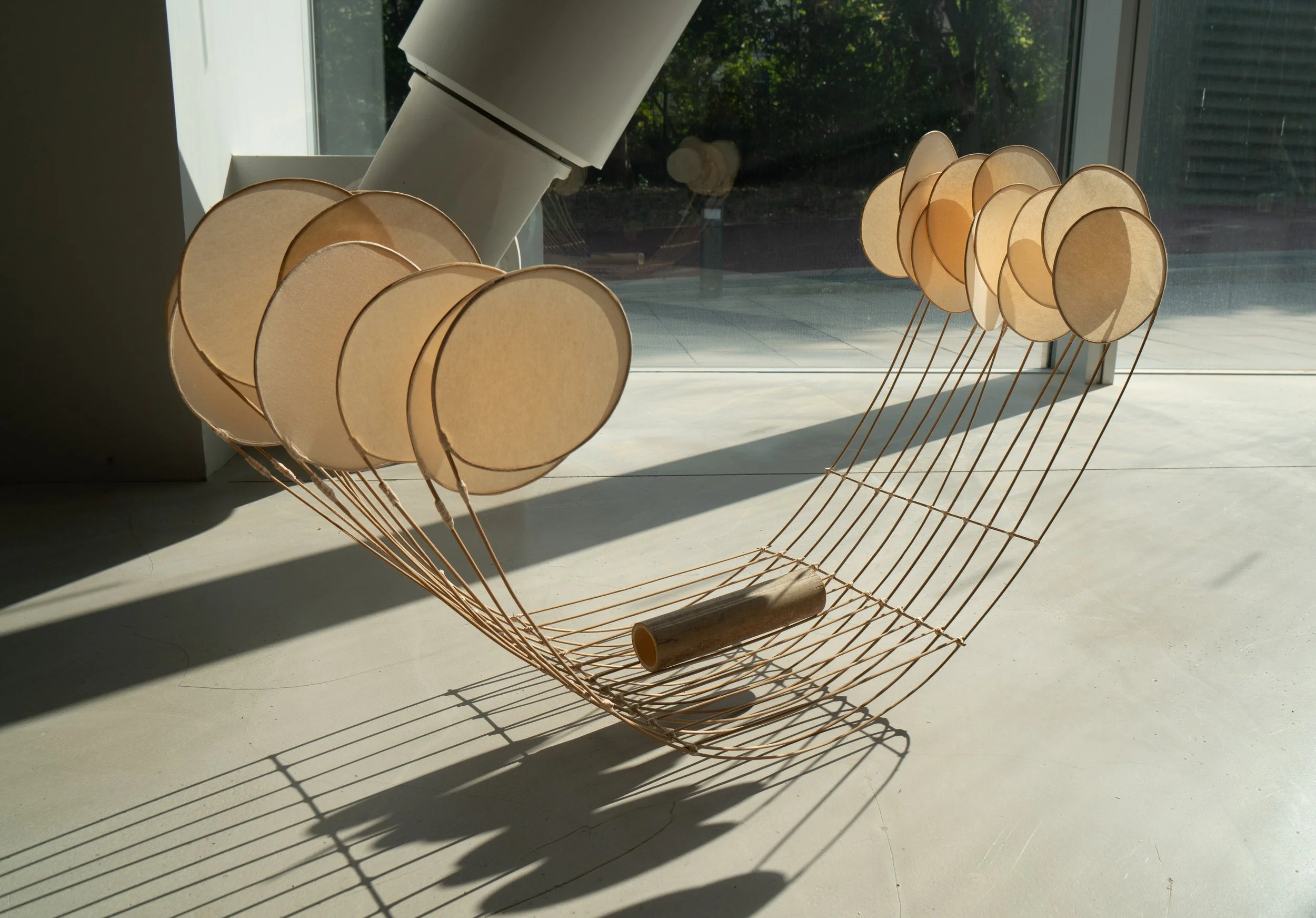 A modern art sculpture resembling a lounge chair made of thin wooden rods with circular cream-colored lampshades attached as the backrest and armrests, placed near a large window with sunlight casting shadows.