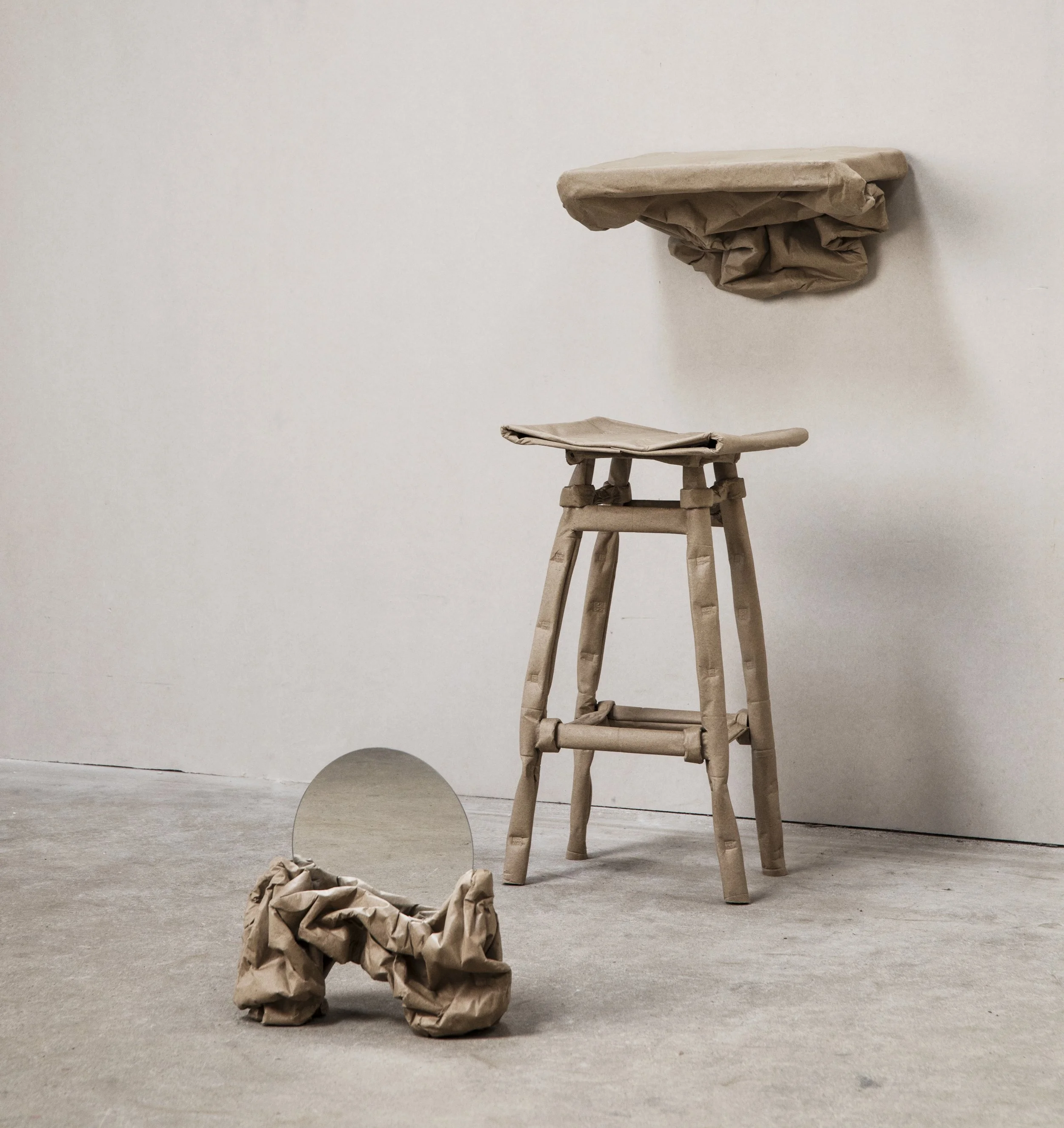 Art installation with crumpled brown paper objects: a wall-mounted object, a small wooden stool, a small circular mirror on the floor, and a crumpled paper object with a mirror leaning against it.
