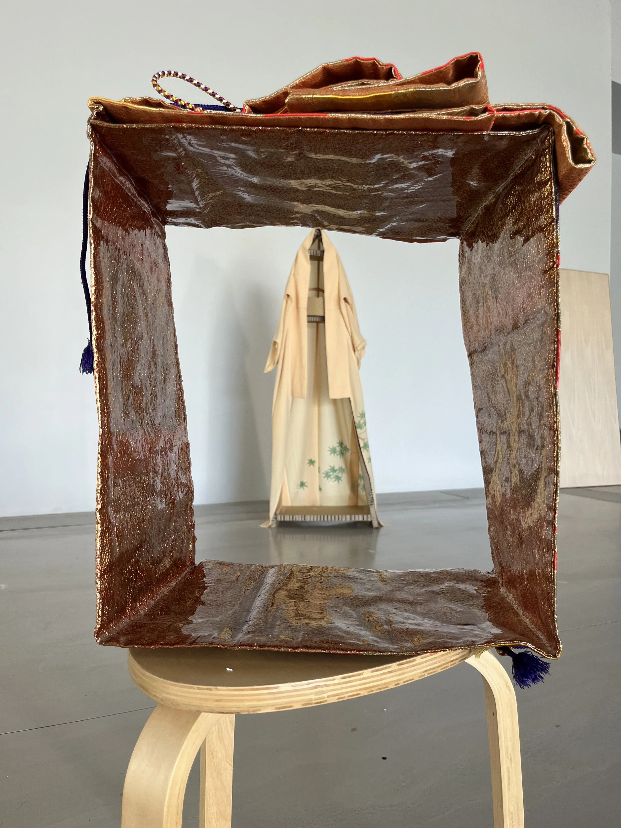 A square-shaped, textured, brown display frame with a glossy finish, framed on a wooden stool, with a mannequin dressed in traditional Japanese kimono in the background.