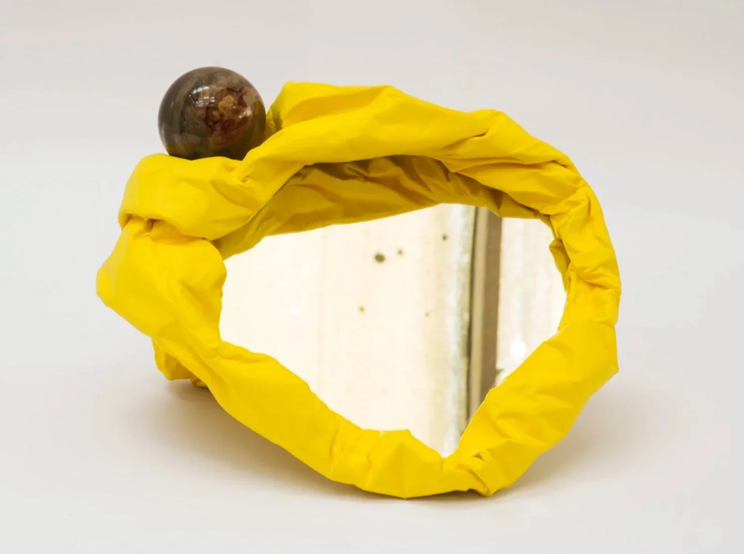 A mirror with a frame made of yellow plastic or paper, with an object on top that appears to be a round, dark-colored fruit or stone.