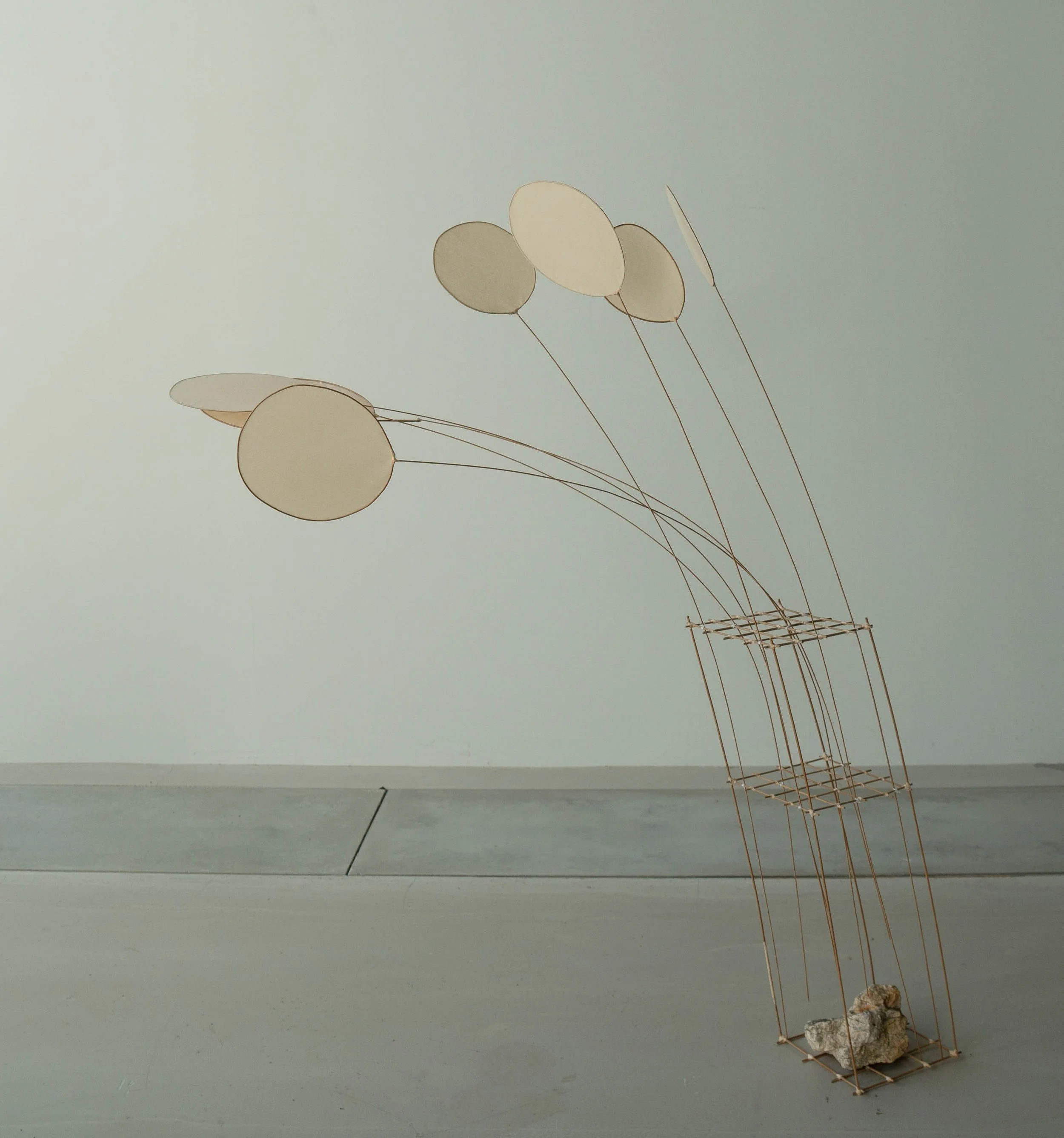 A minimalist sculpture of a tower with a base of rocks and metal rods, with metal rods extending upward to hold large oval-shaped discs resembling balloons in neutral tones.