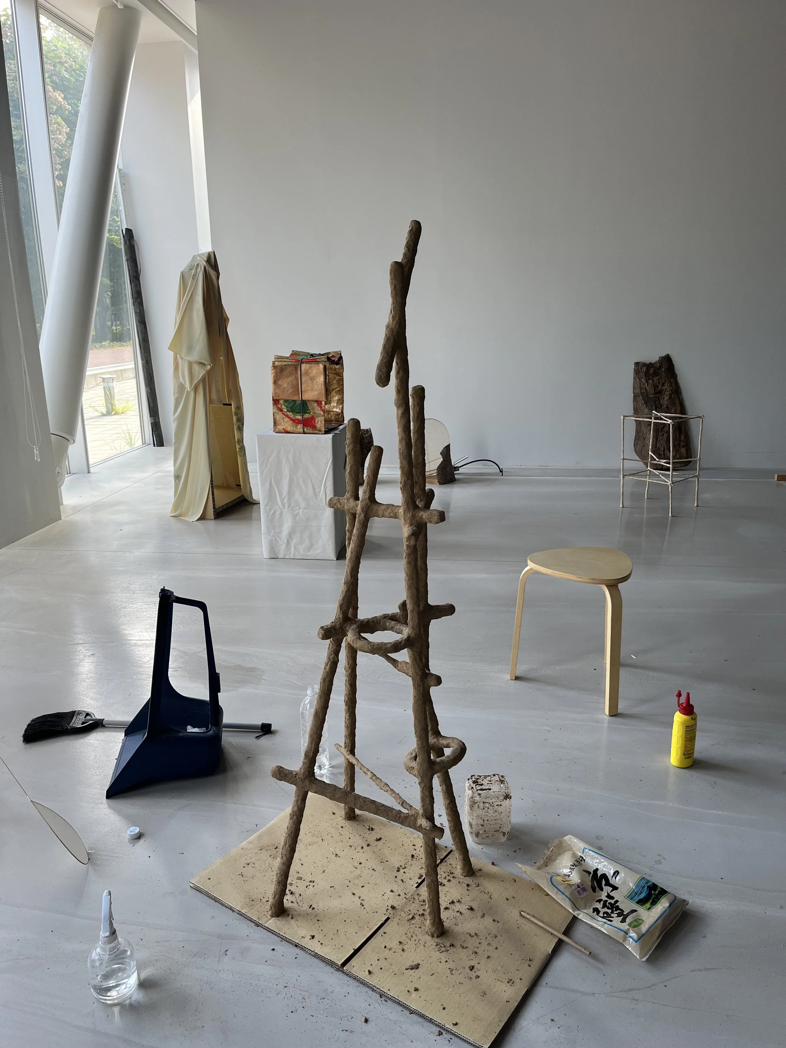 Artist's workspace with a stick sculpture in progress, surrounded by tools, a small stool, and various art supplies, in a modern, well-lit gallery space.