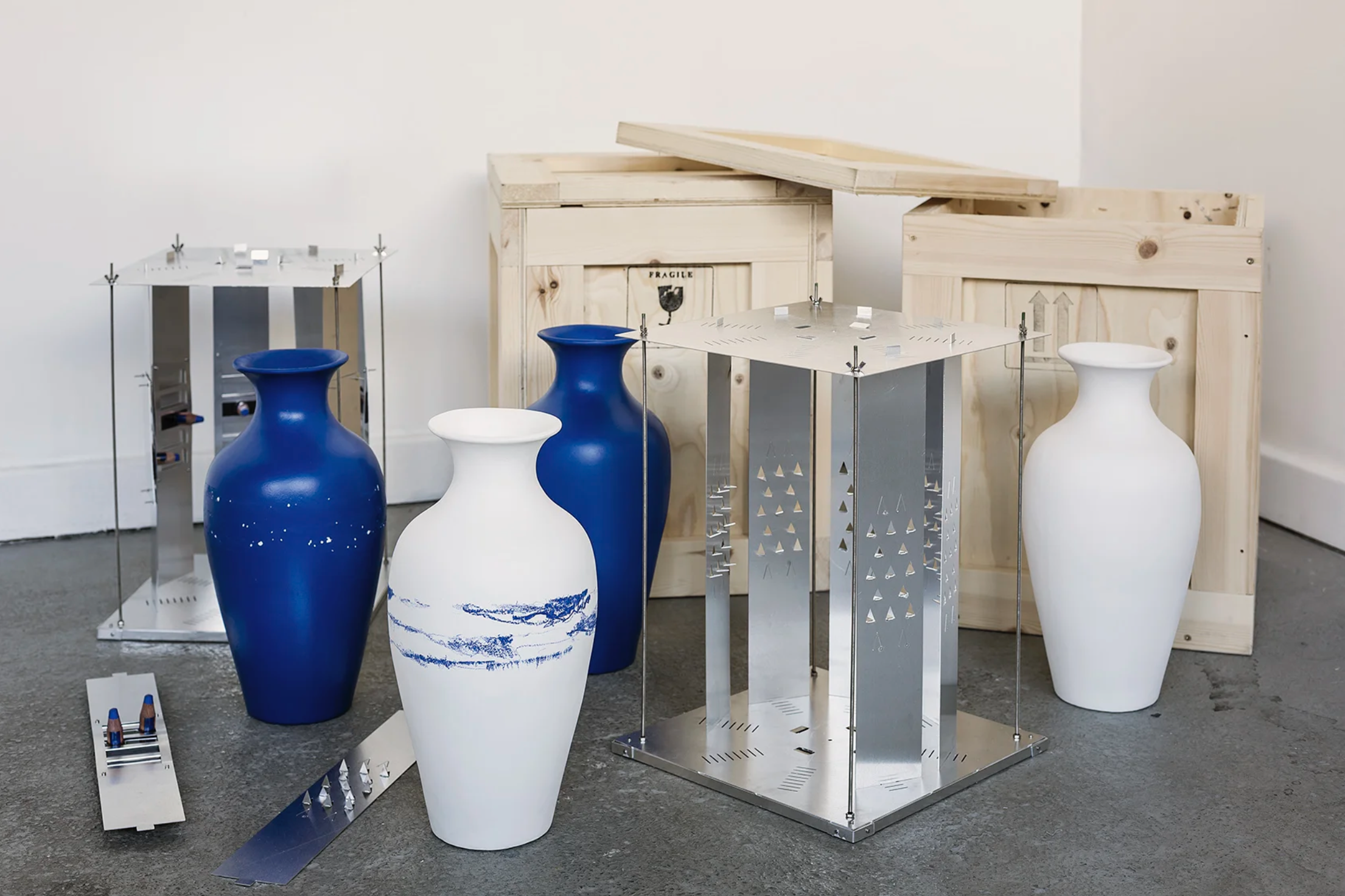 Four large vases, two white with blue patterns and two solid blue, are arranged among metal frames, wooden crates, and metal panels in a gallery setting.