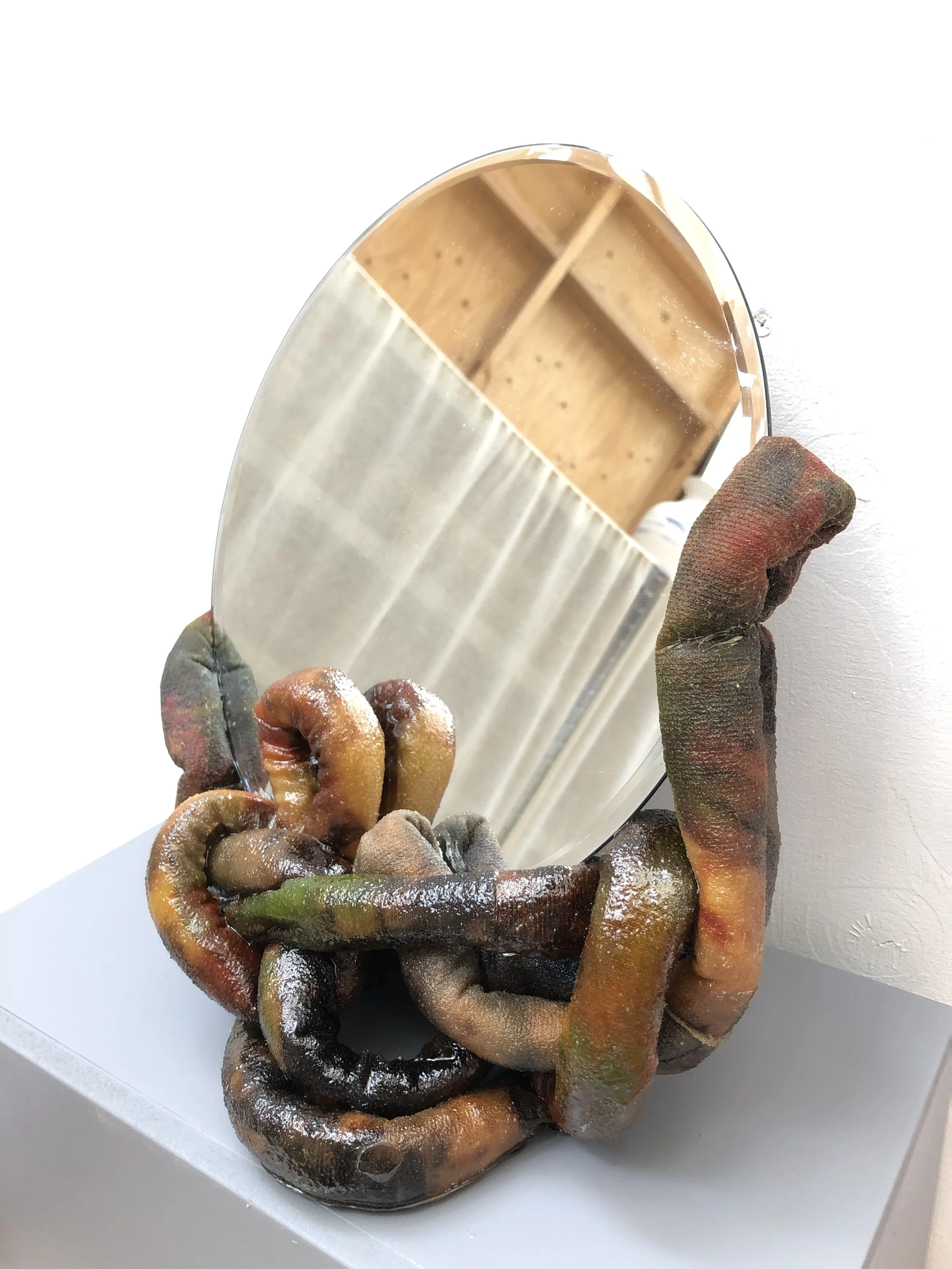A mirror with a frame made of intertwined, multicolored, rusty and decayed-looking rubber tubes or coils, placed on a white surface against a white wall. The reflection shows a wooden wall with a curtain and a ceiling light.