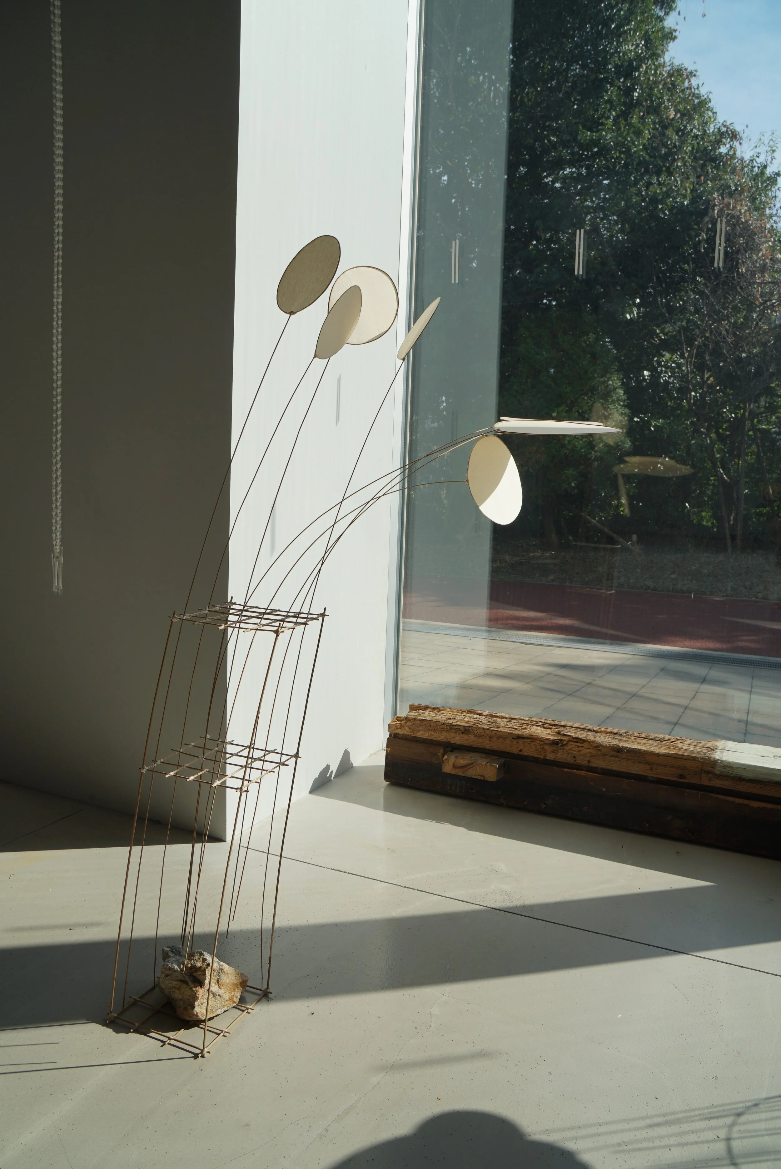 Metal sculpture with oval shapes in front of a glass window, with sunlight casting shadows on a white floor.