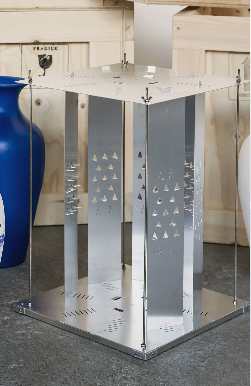 Metal structure with perforated panels and four vertical rods, resting on a concrete floor in front of a wooden wall, with a large blue ceramic vase on the left.
