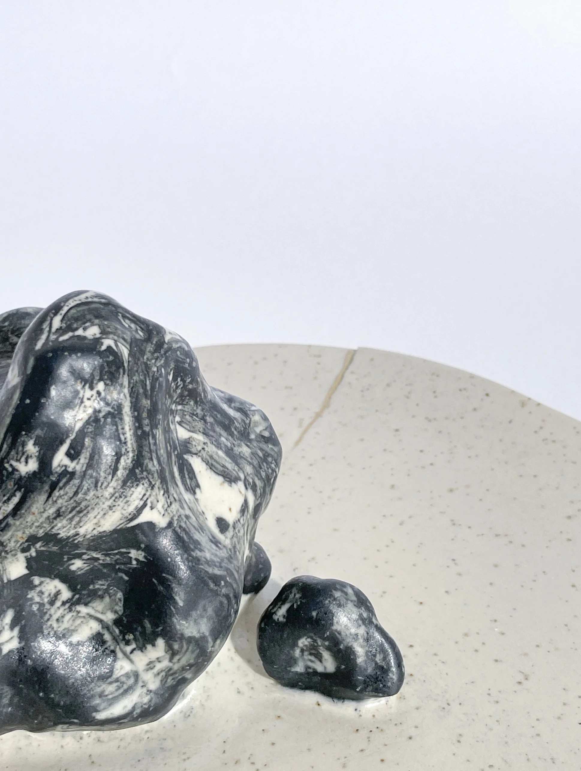 Close-up of a marbled black and white ceramic sculpture resembling a dog, with a small black stone beside it on a white speckled surface, against a plain white background.