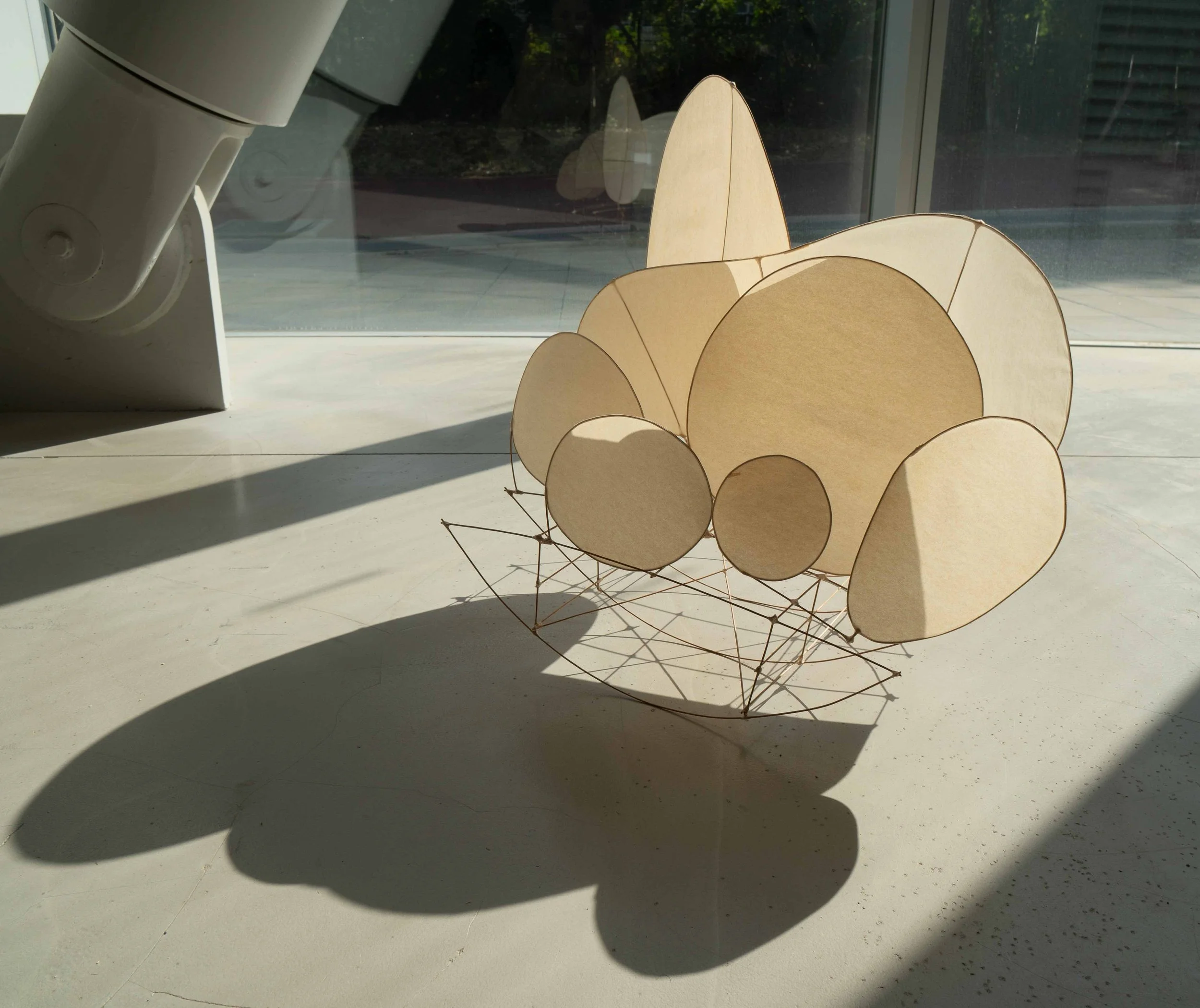 A modern sculpture resembling a flower or abstract shape made of beige wooden circles and petal-like pieces, with thin metal rods forming its base, situated on a stone surface with shadows cast by sunlight.
