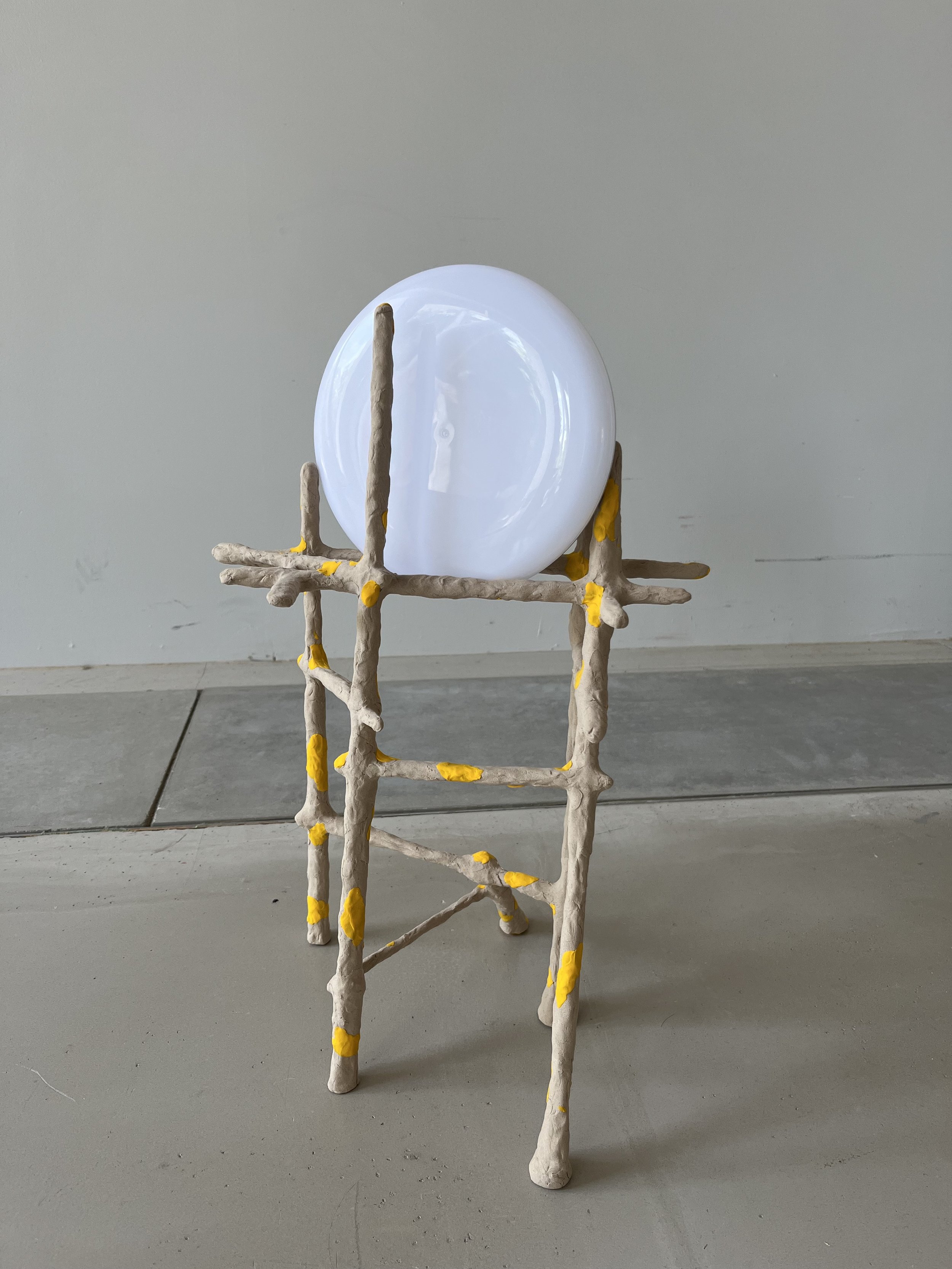 A white plate balanced on a sculpture made of beige sticks with yellow accents, arranged in a ladder-like structure, on a gray tiled floor against a plain light-colored wall.