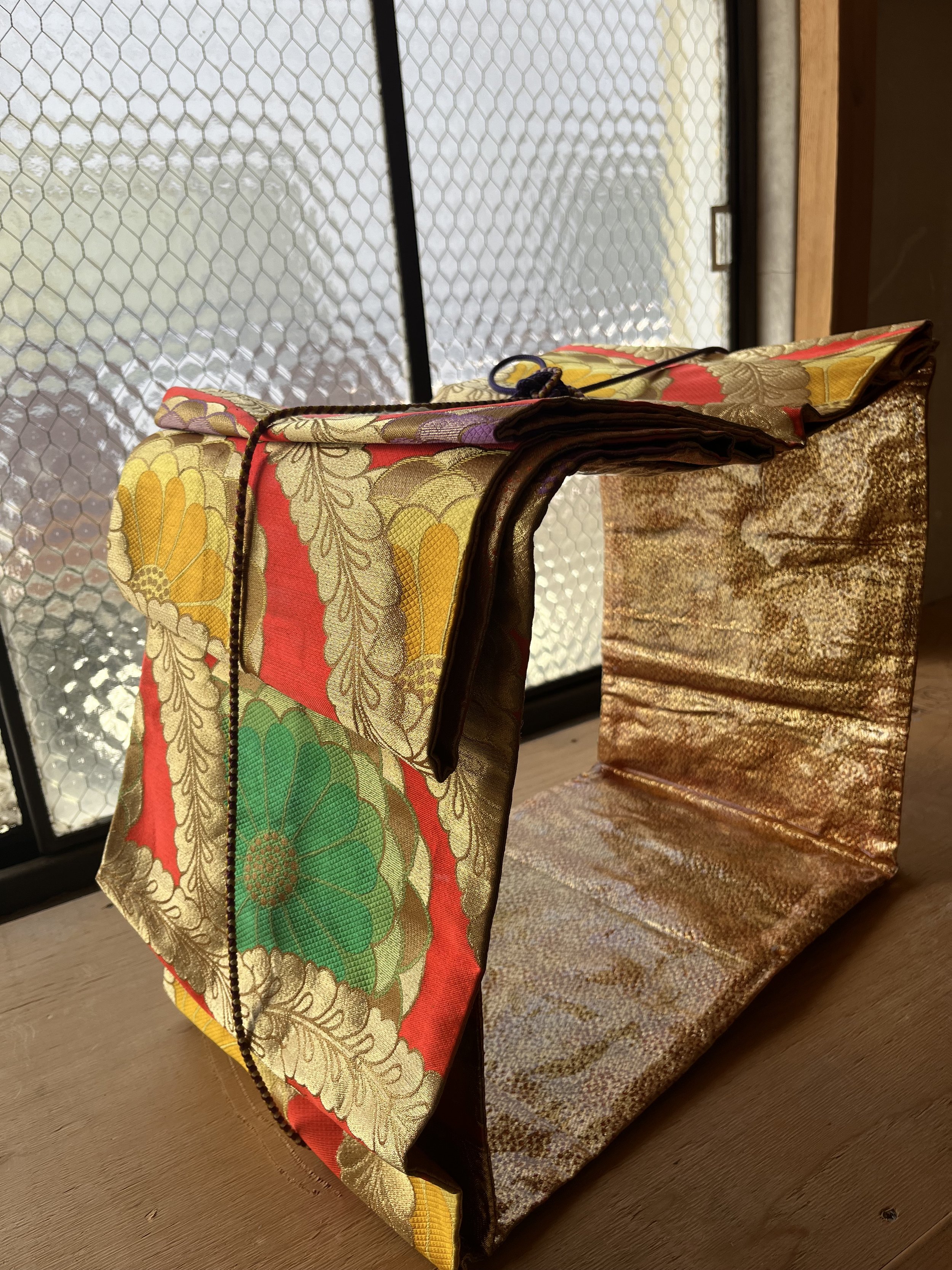 A colorful, ornate handbag made of fabric with gold, green, yellow, red, and purple floral patterns sits on a wooden surface near a window with textured glass.