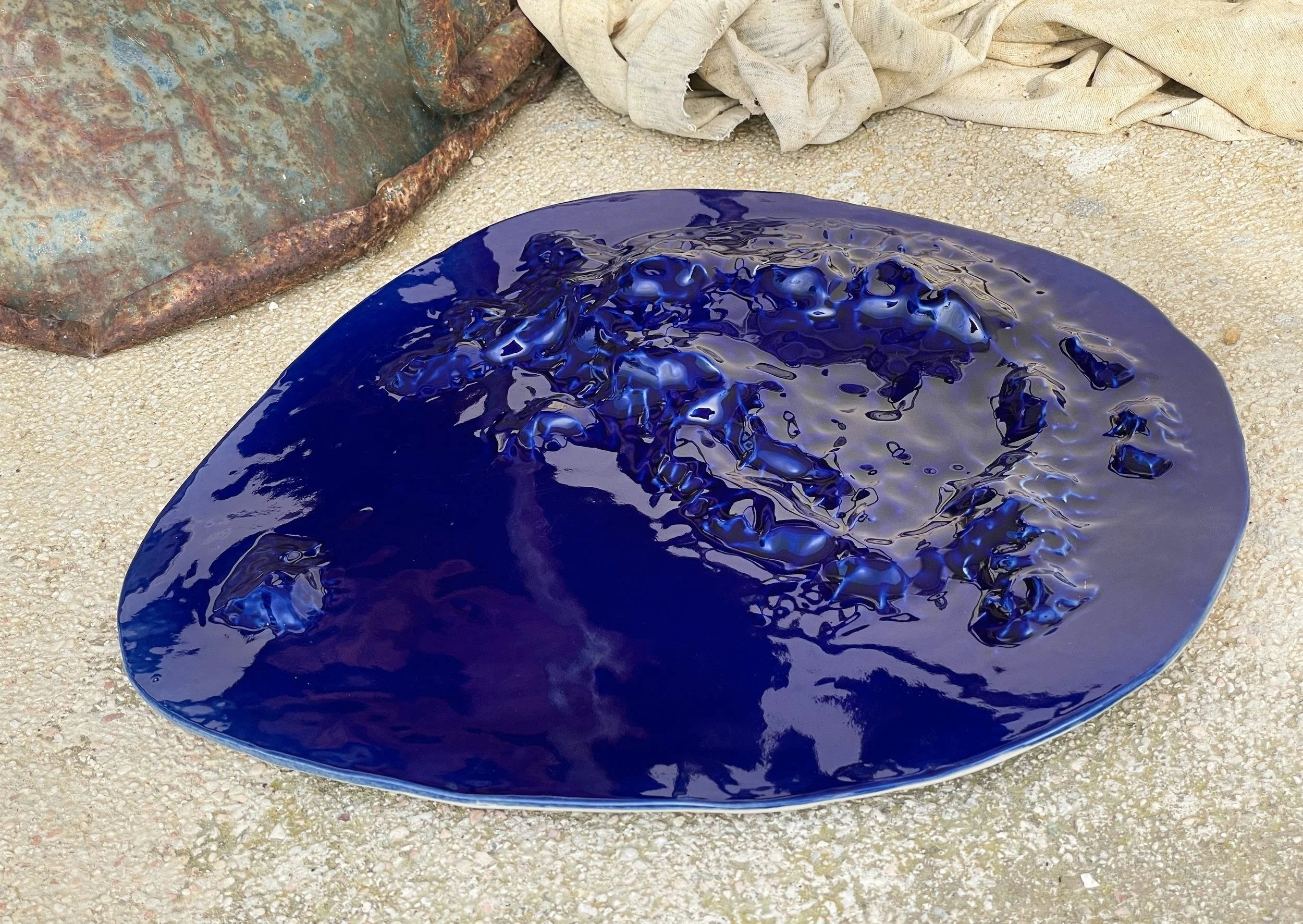 A large, irregularly shaped, glossy blue ceramic plate with textured, uneven surface, resting on a sandy ground with a rusted metal object and some fabric in the background.